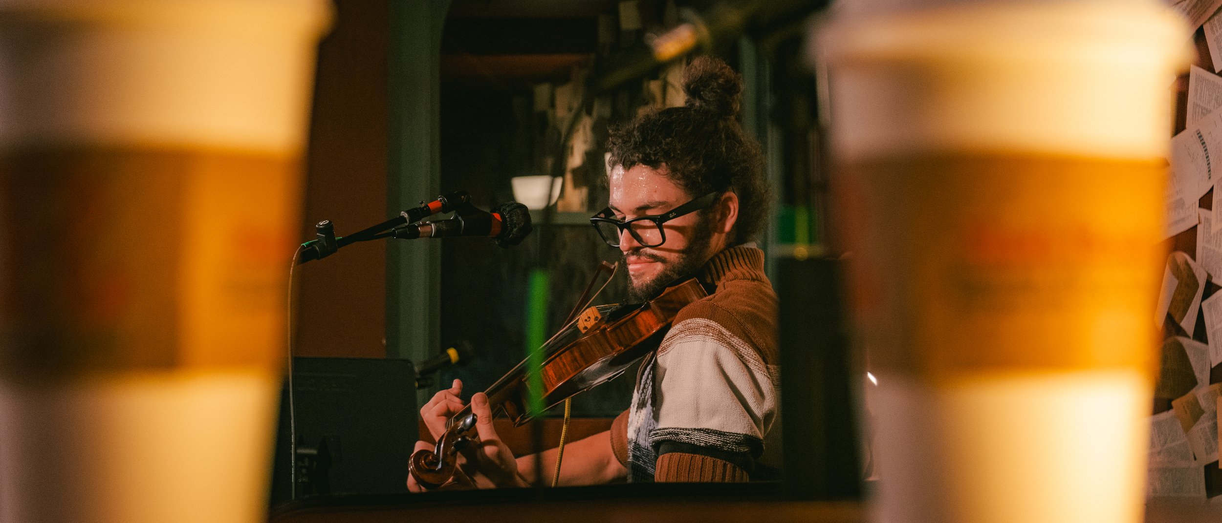 A man with glasses and a beard playing a violin in a music studio, seen through a mirror or window with two large coffee cups in the foreground.