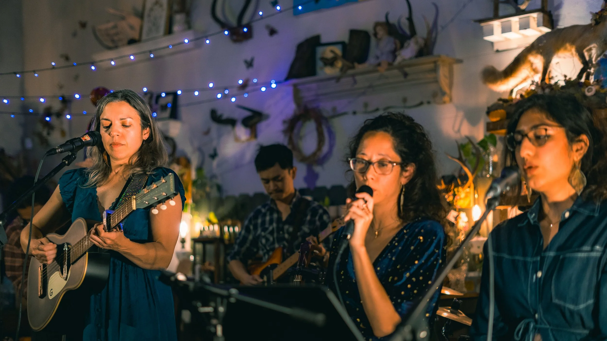 Women performing music indoors, with one playing an acoustic guitar and others singing, decorated with string lights and wall ornaments.
