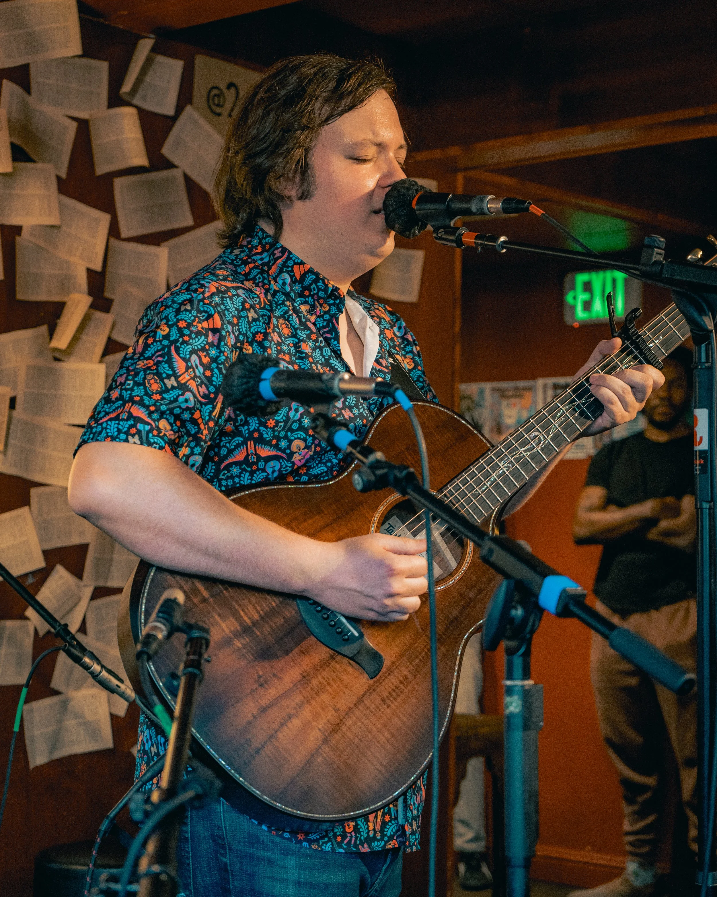 A person singing into a microphone while playing an acoustic guitar in a dimly lit room with papers on the wall and a man standing in the background.
