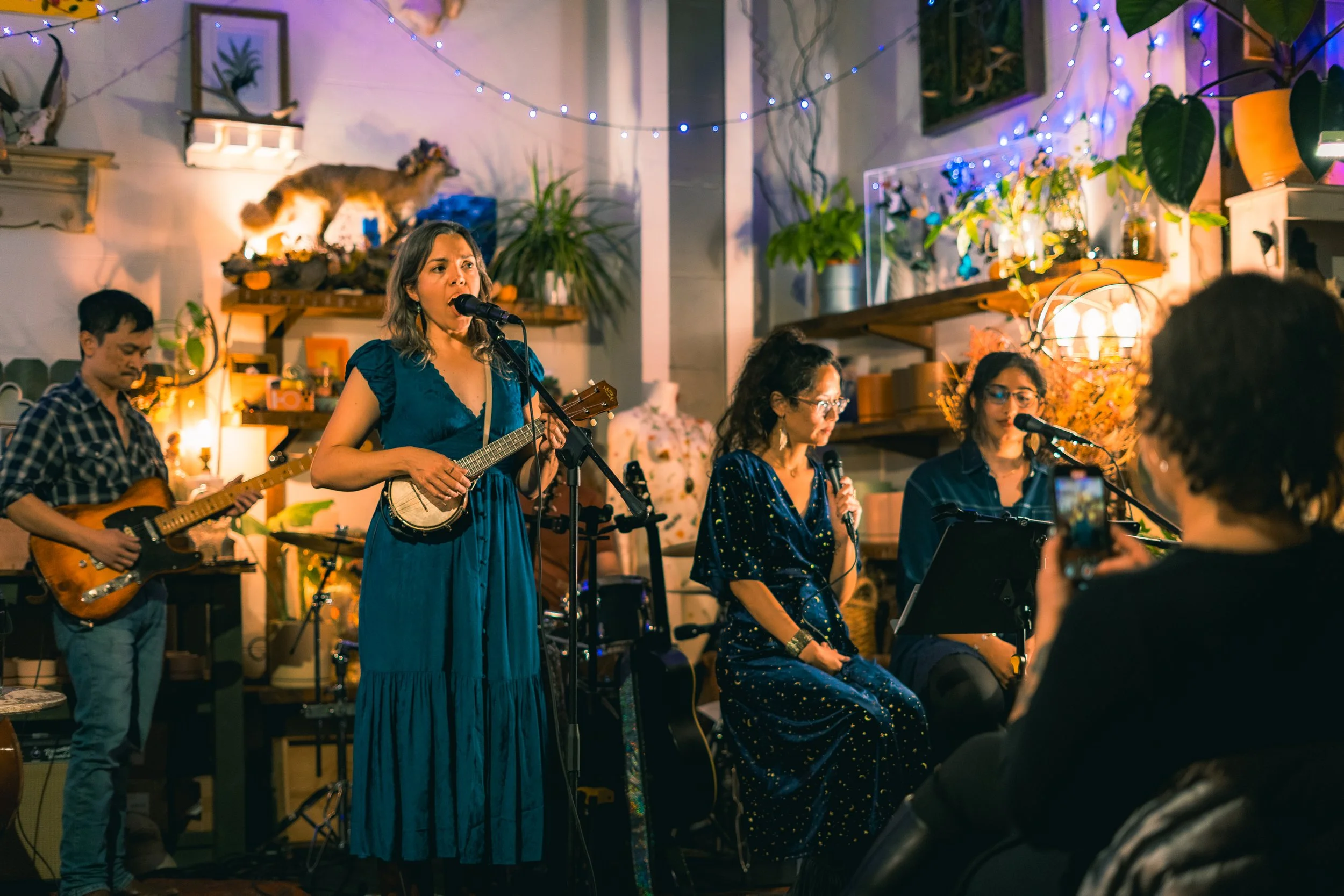 A group of four women and a man performing music together in a cozy, decorated indoor space with string lights, plants, and artwork. One woman is singing into a microphone and playing a ukulele, while another woman is sitting, singing, and wearing gl