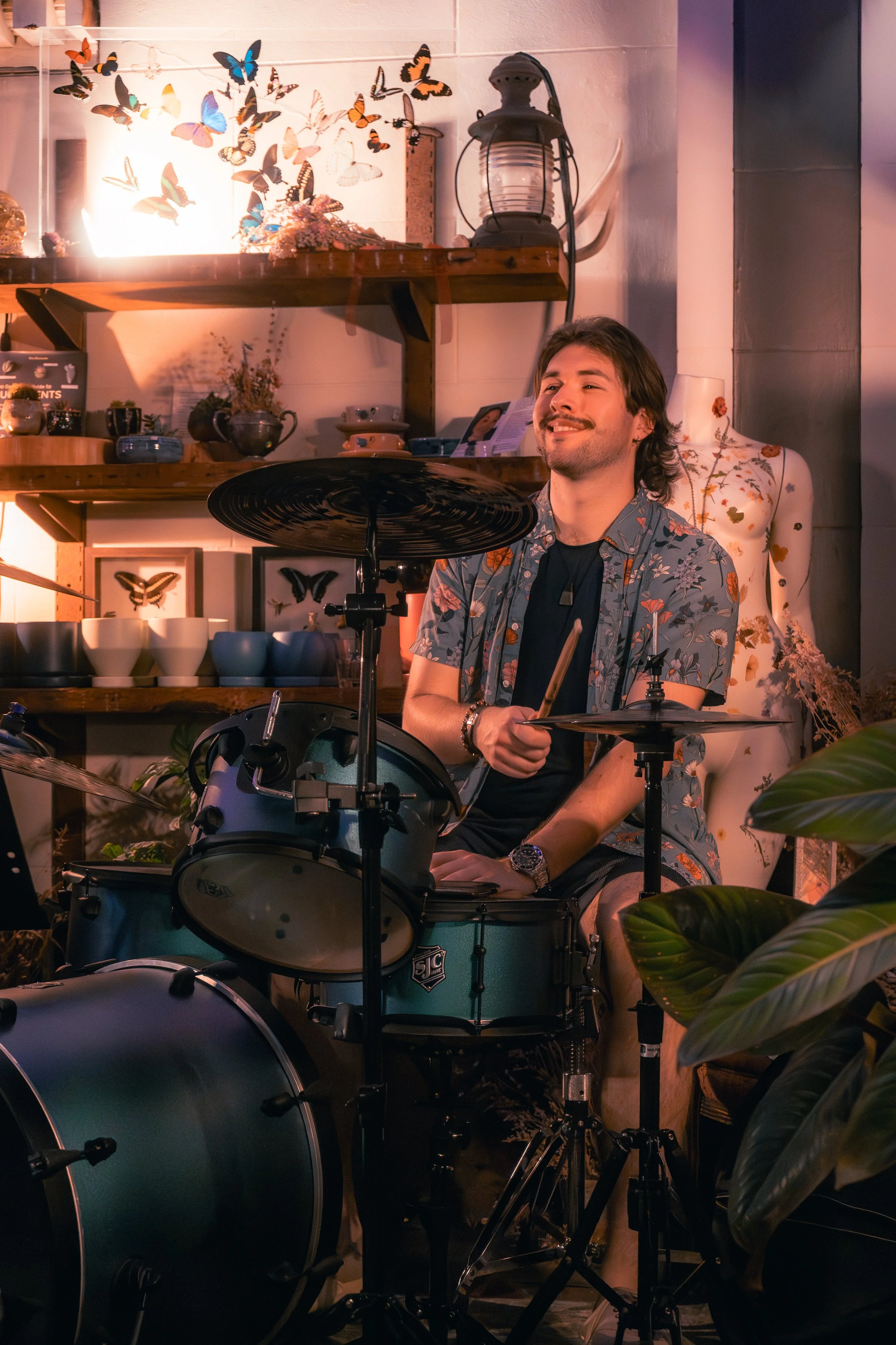 A man playing the drums indoors, surrounded by decorative items including butterfly wall art, shelves with teapots and bowls, and a mannequin with a floral pattern. The setting has warm lighting and a cozy atmosphere.
