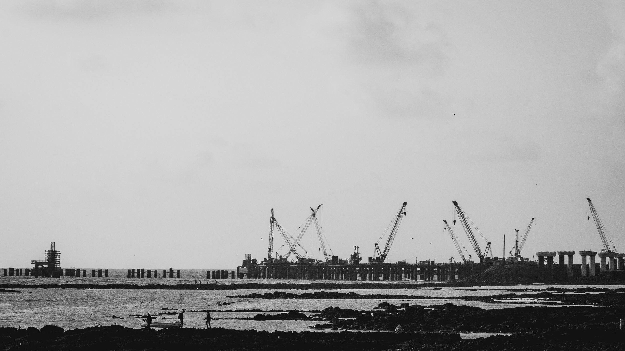 Black and white photograph of a construction pier with cranes, distant ocean, and people on rocky shoreline.