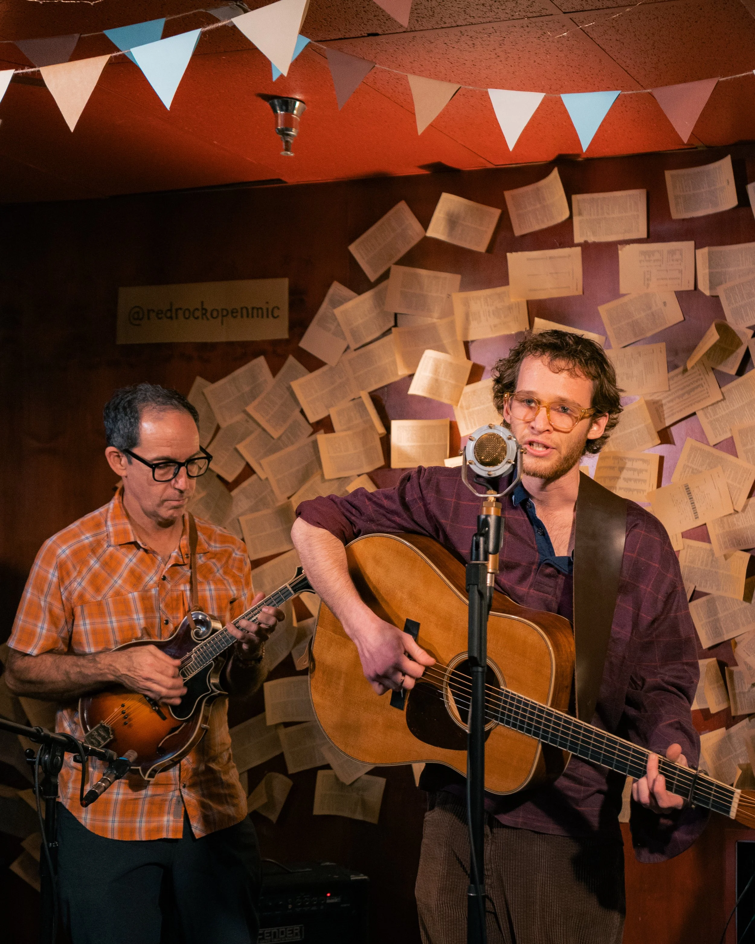 Two musicians perform on stage, one playing a mandolin and the other playing an acoustic guitar. The background features a wall decorated with open books and string lights, with a sign that reads '@redrockopenmic'.