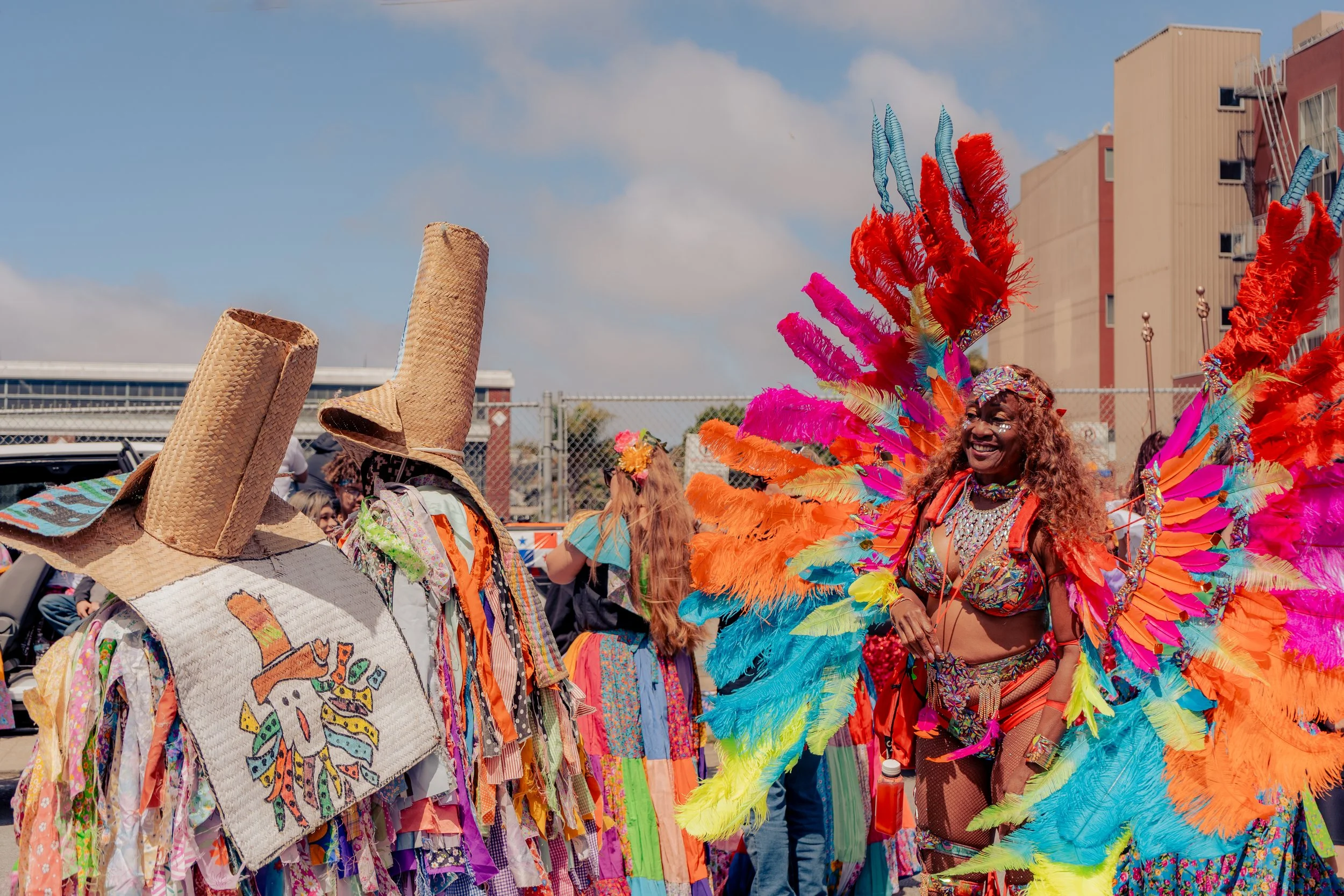 Colorful parade with a woman in a vibrant feathered costume and other people in elaborate costumes and large straw hats near a fence and buildings.