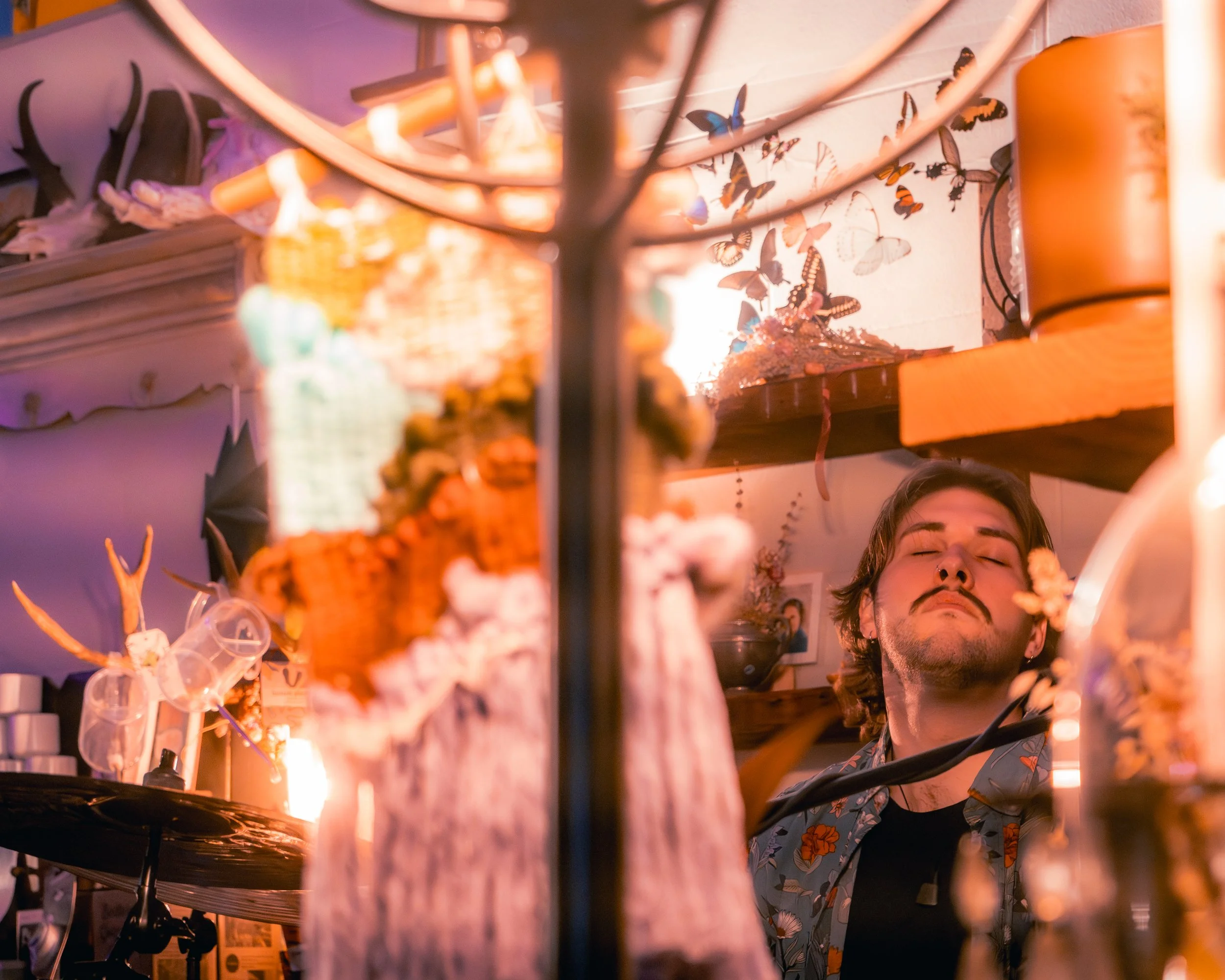 A man with closed eyes and a relaxed expression, standing behind a bar or counter decorated with flowers and butterflies, in a warm, ambient light.