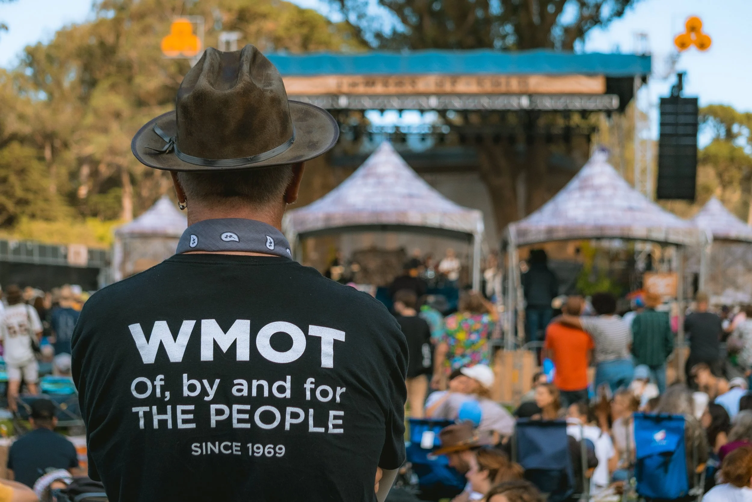A person wearing a brown cowboy hat and a black T-shirt with the words 'WMOT Of, by and for THE PEOPLE SINCE 1969' on the back is watching a crowd gathered at an outdoor concert stage with a blue roof and tents.