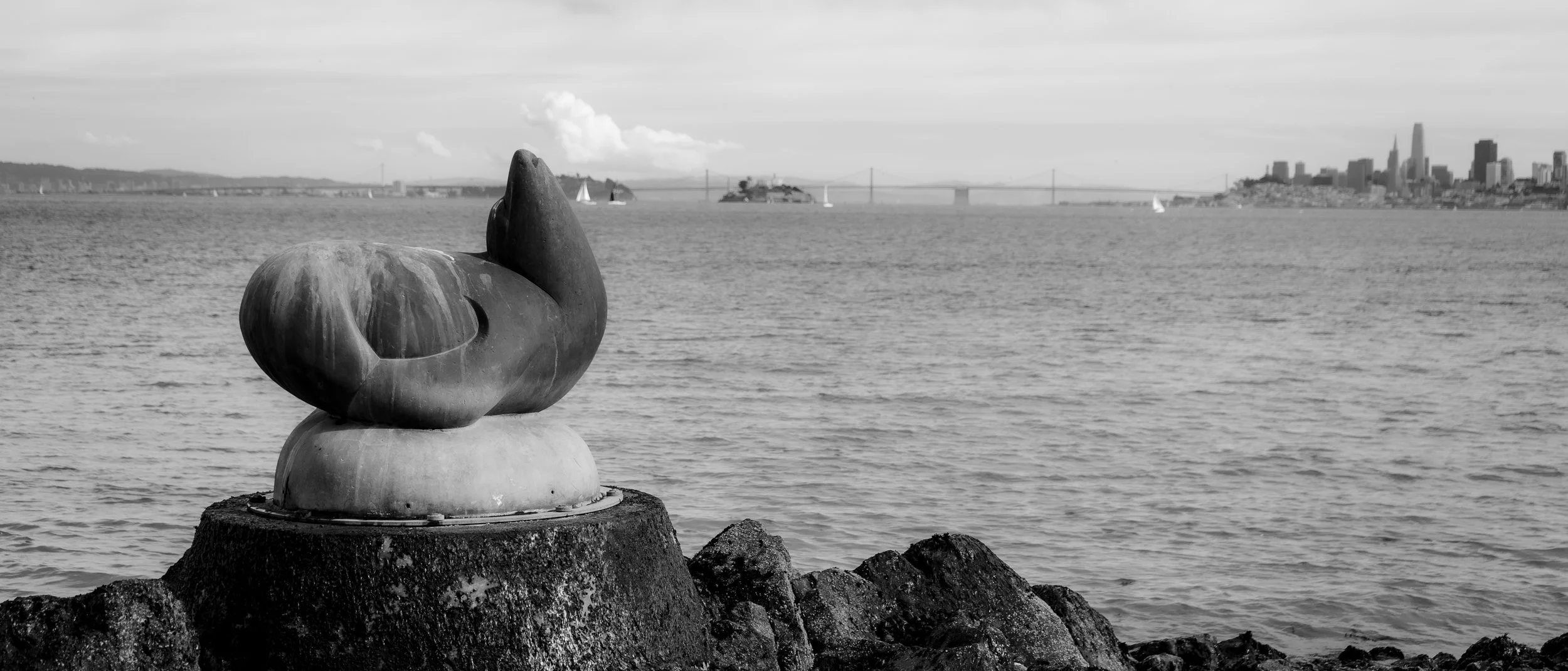 A stone sculpture on the water in Sausalito with the SF city skyline and Bay Bridge in the background, in black and white.