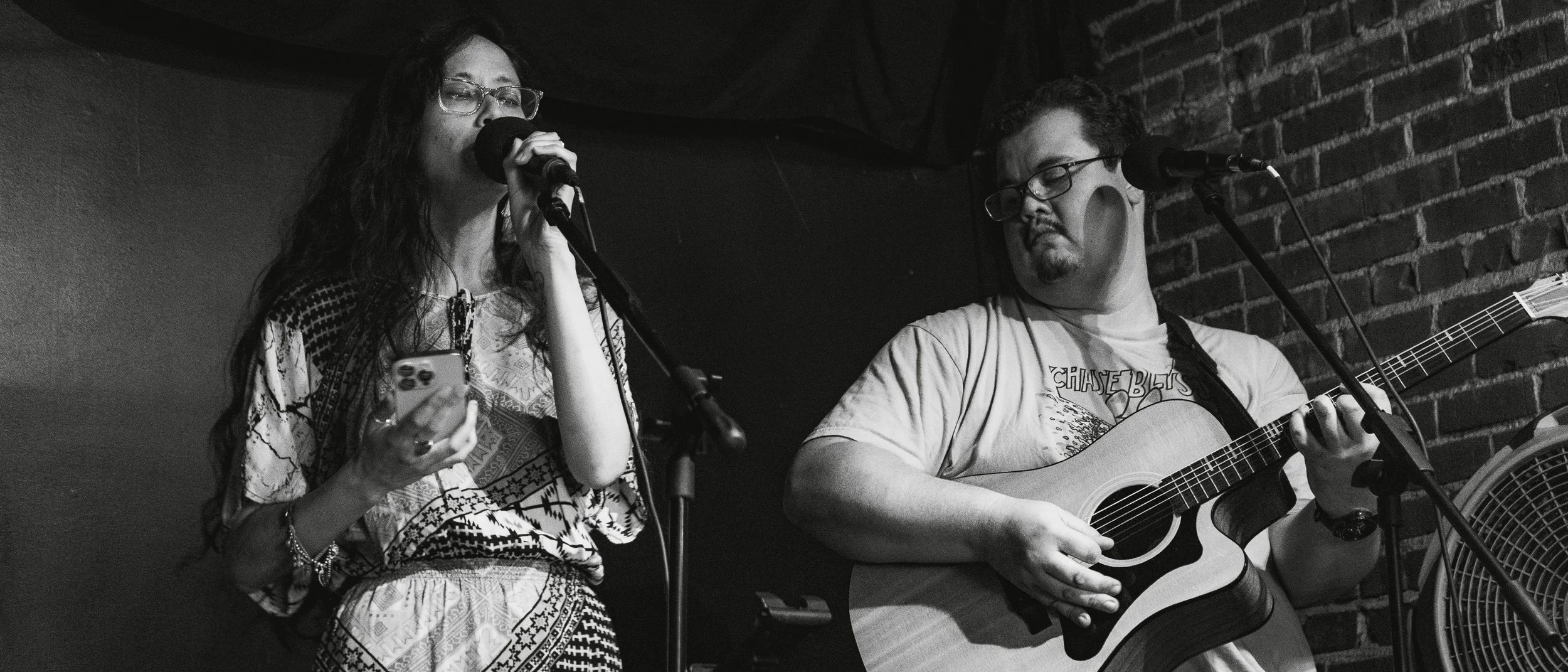 A woman singing into a microphone while looking at her phone, and a man playing an acoustic guitar on a brick wall stage in black and white.