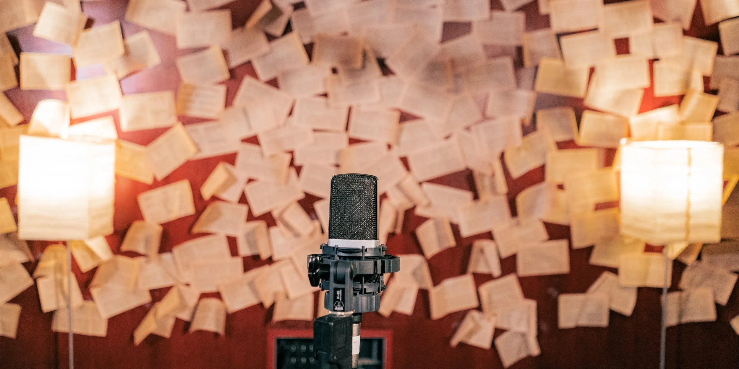A microphone on a stand positioned in front of a wall decorated with open books, with two lamps on either side casting warm light.