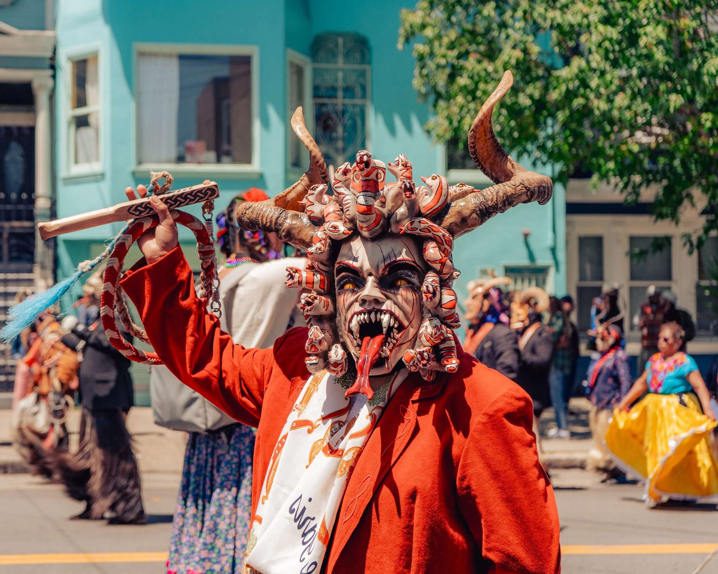 Person dressed as the Mexican holiday figure La Catrina, wearing an elaborate mask with snakes, horns, and a devil's face, holding a stick, during a parade with colorful costumes and a crowd in the background.