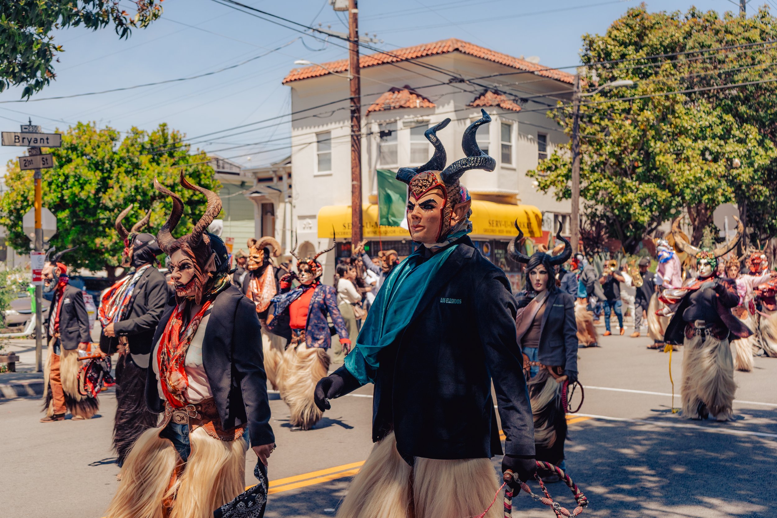 People dressed in traditional Mexican costumes with horns, masks, and long skirts participating in a street parade on a sunny day.
