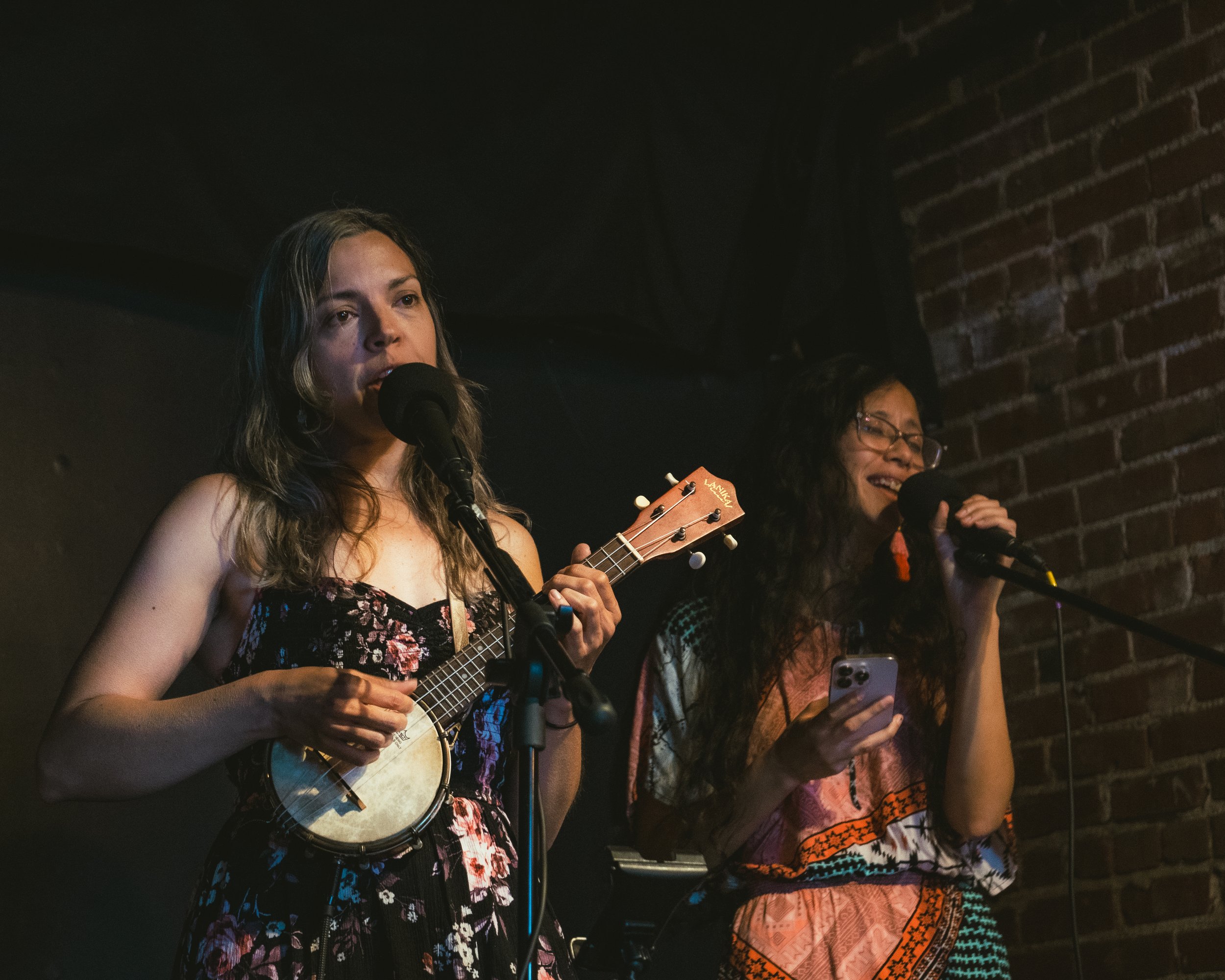 Two women singing into microphones, one playing a ukulele, in a dimly lit venue with a brick wall background.