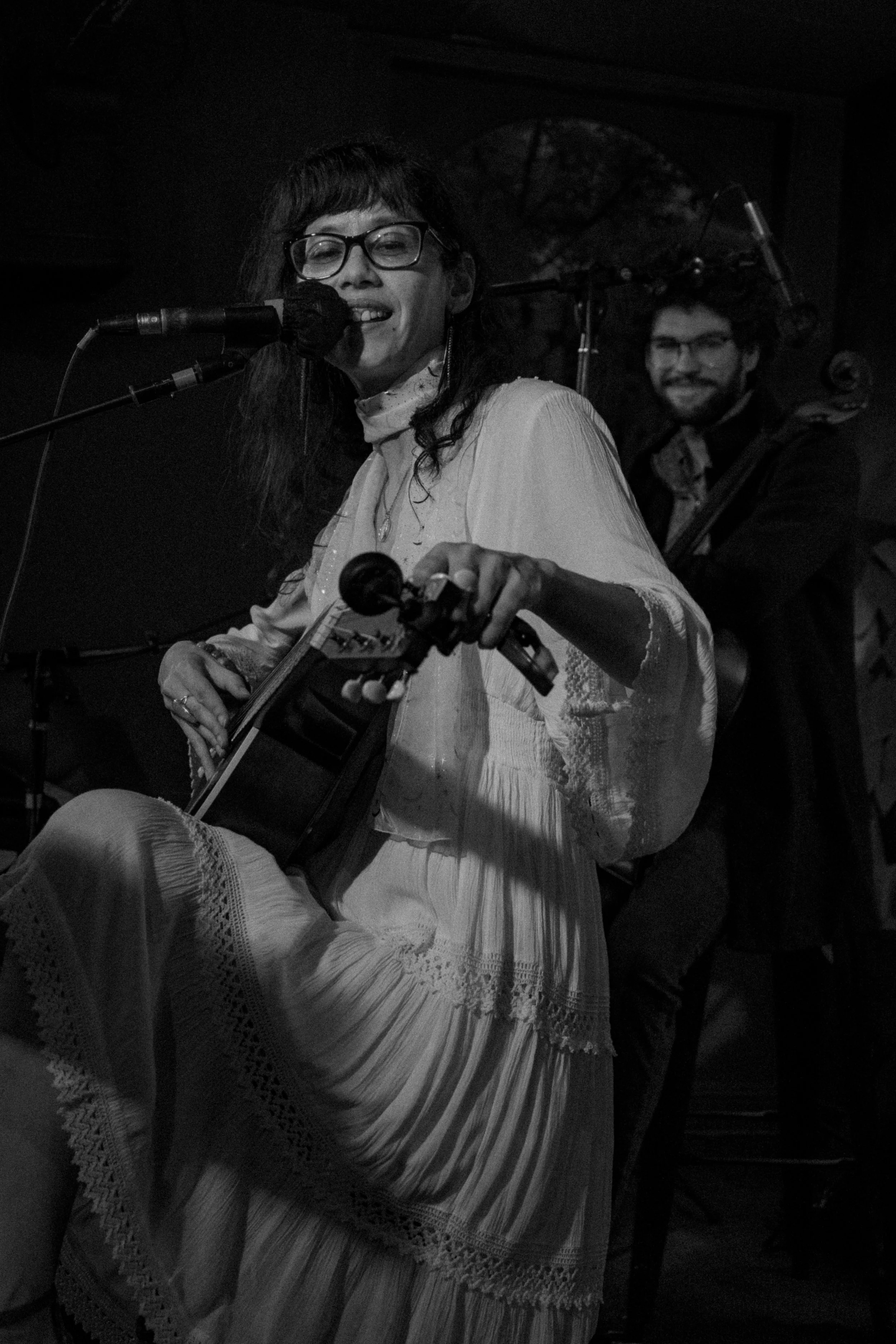 A woman in a white dress playing an acoustic guitar and singing into a microphone with a man in the background playing a double bass in a dimly lit setting.