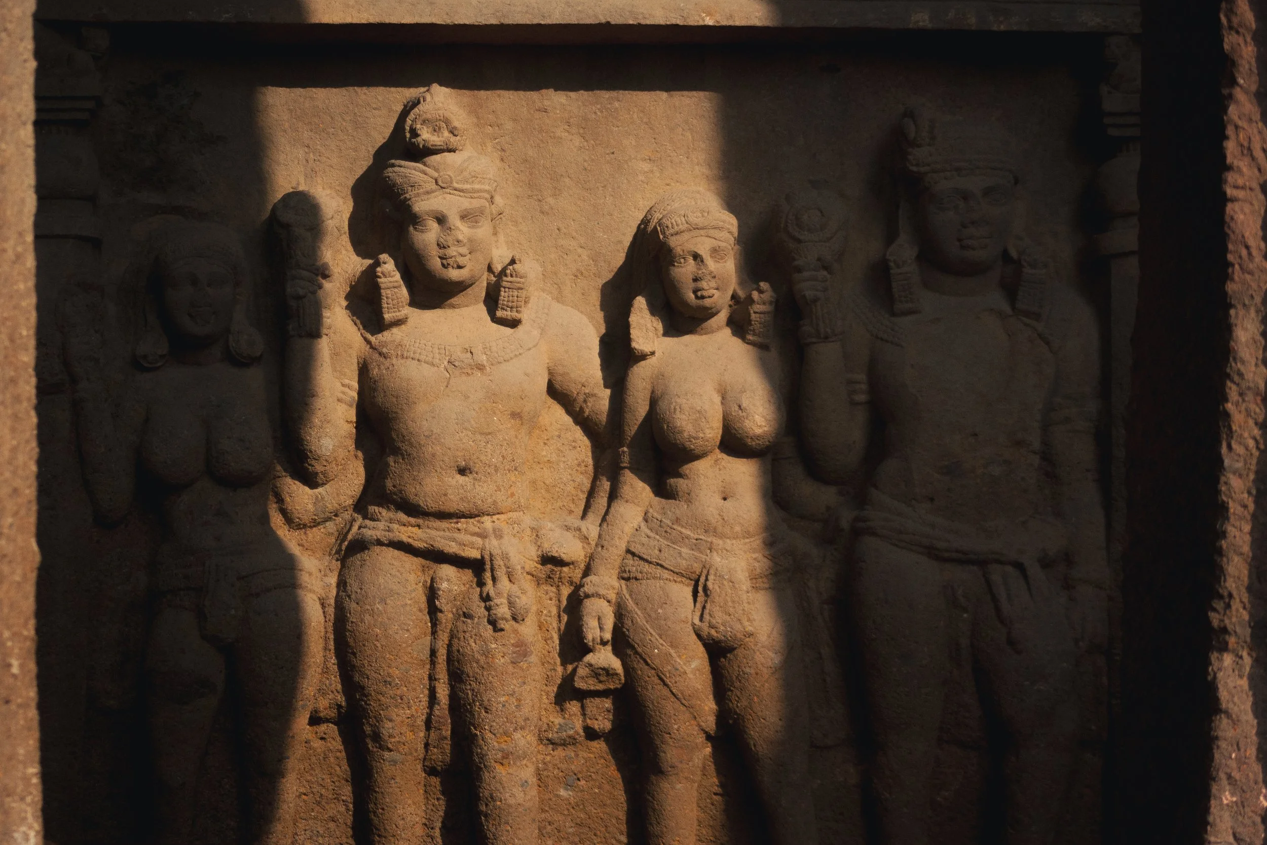 Ancient stone carvings of four figures with intricate jewelry, headdresses, and clothing, illuminated by sunlight, in a historical archaeological context.