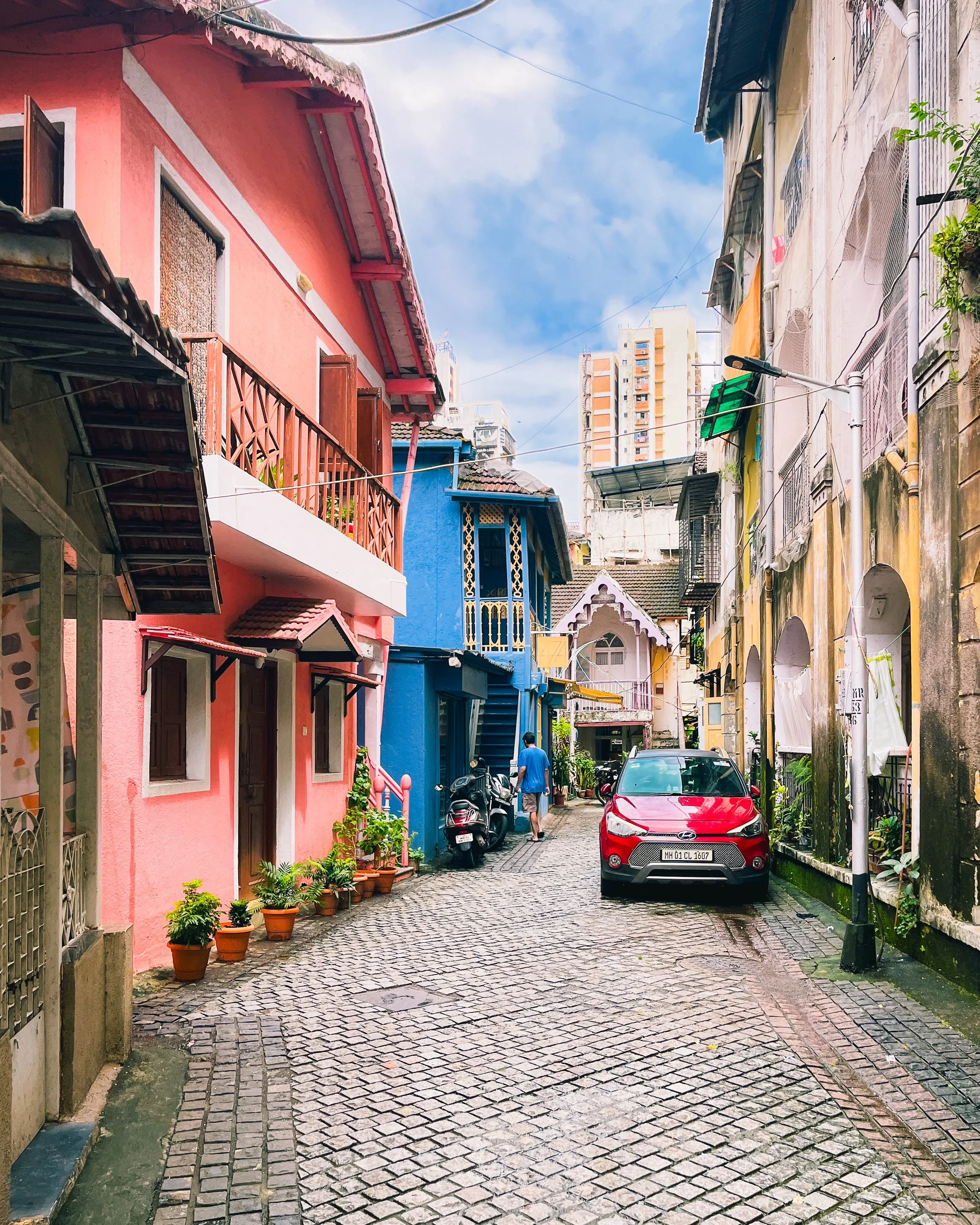 Colorful neighborhood street with pink, blue, yellow, and white houses, small potted plants, a red car, scooters, and a person walking down the cobblestone street under a partly cloudy sky.