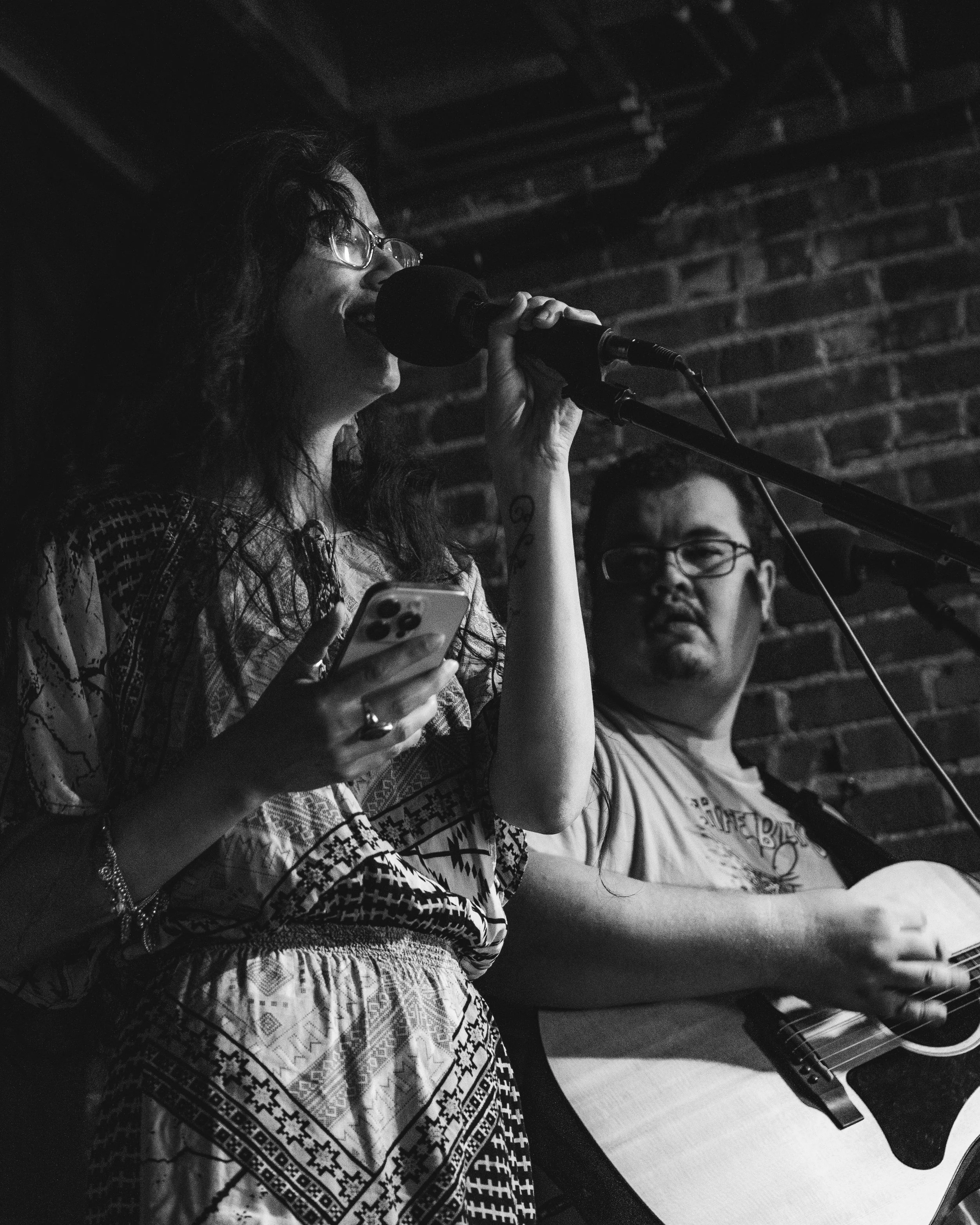 A black-and-white photo of a woman singing into a microphone while holding a phone, and a man playing an acoustic guitar against a brick wall background.