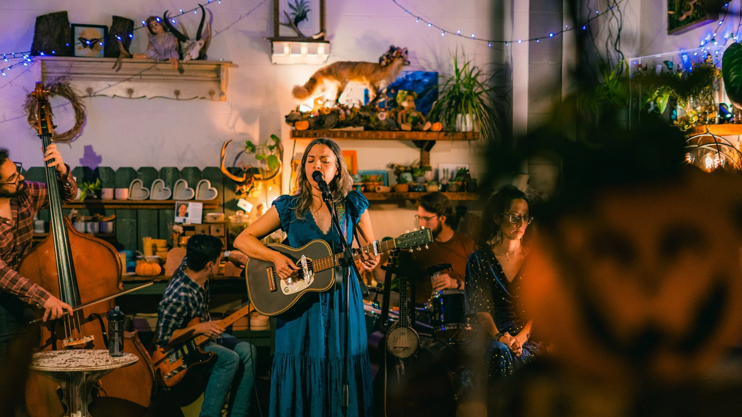 A woman singing and playing guitar in a cozy, decorated indoor setting with other musicians, fairy lights, and various decorations such as a mounted cat and plants.