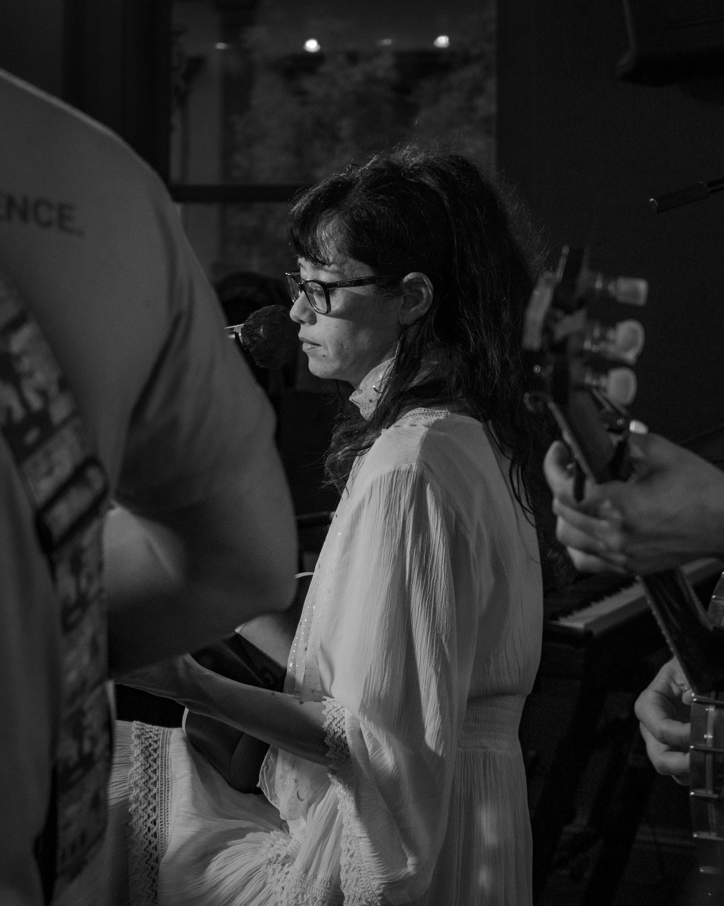 A woman wearing glasses and a white dress is singing into a microphone while playing a piano. She is surrounded by musicians playing guitars. The scene is in a dimly lit room.