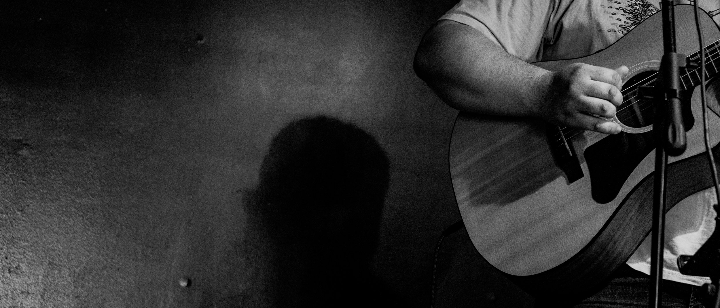 A person playing an acoustic guitar in a recording studio with a microphone in front of the guitar, casting a shadow on the textured wall.