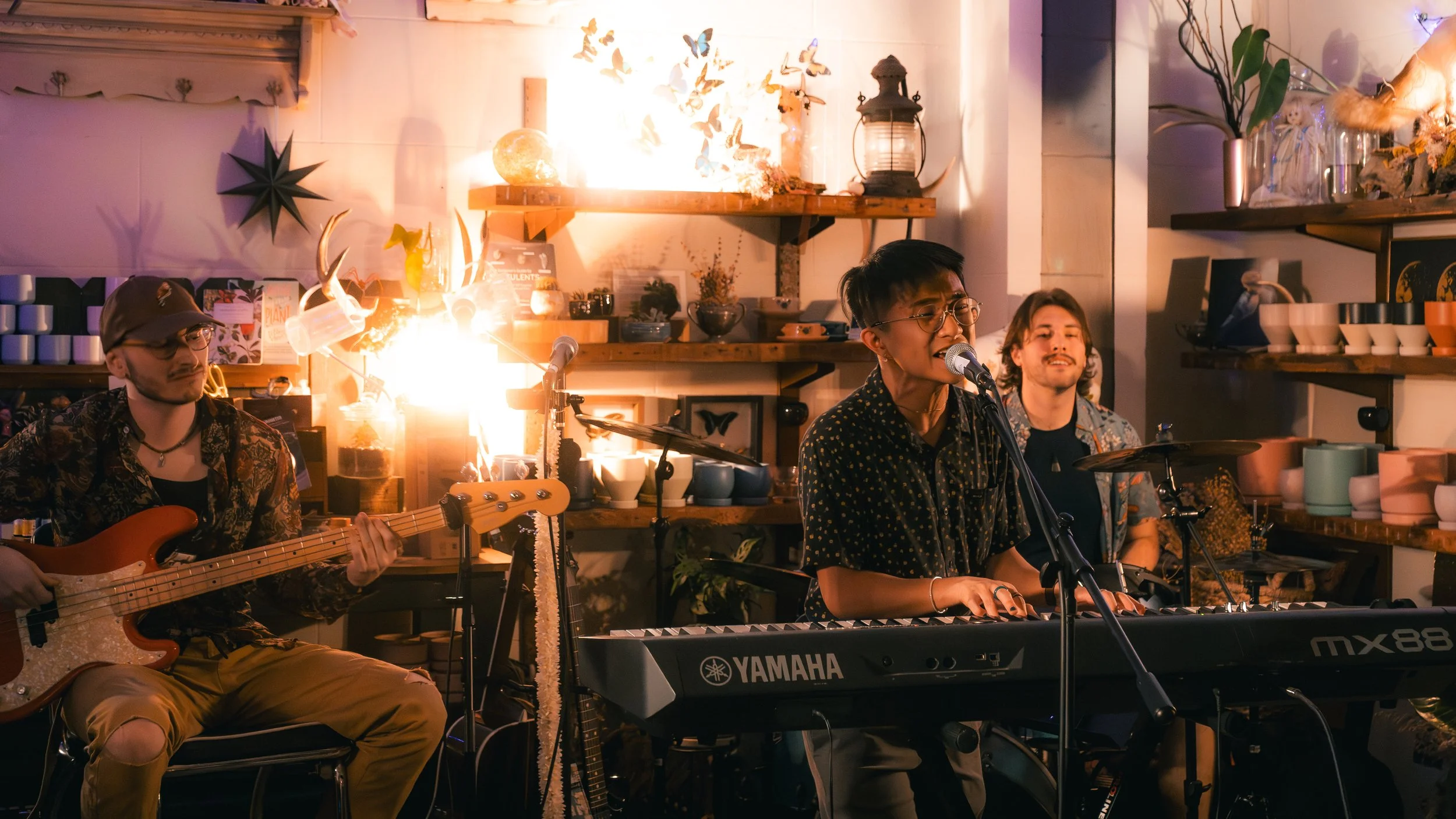 A band performs live with a guitarist, a keyboardist singing into a microphone, and a drummer. The setting is cozy with warm lighting, shelves with ceramics and decorations, and a bright fire or light source behind them.