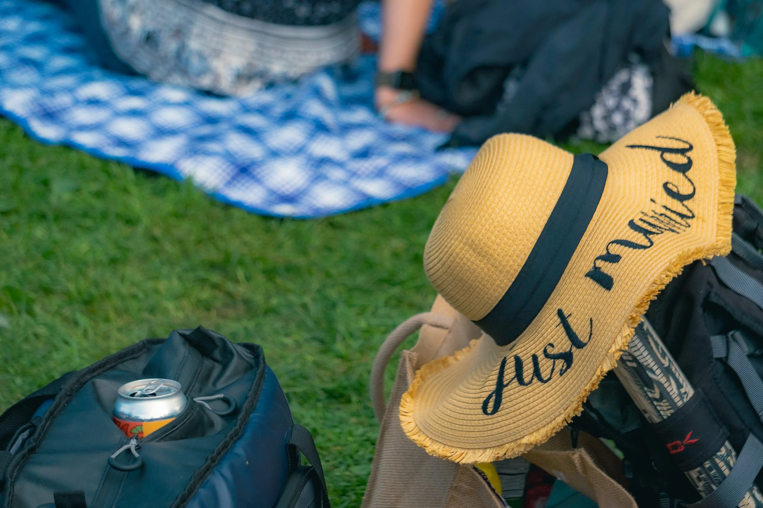 A yellow straw sun hat with black writing that says "just relax" on it, resting on a backpack outside on the grass near cans of drink, with a picnic blanket and bag in the background.