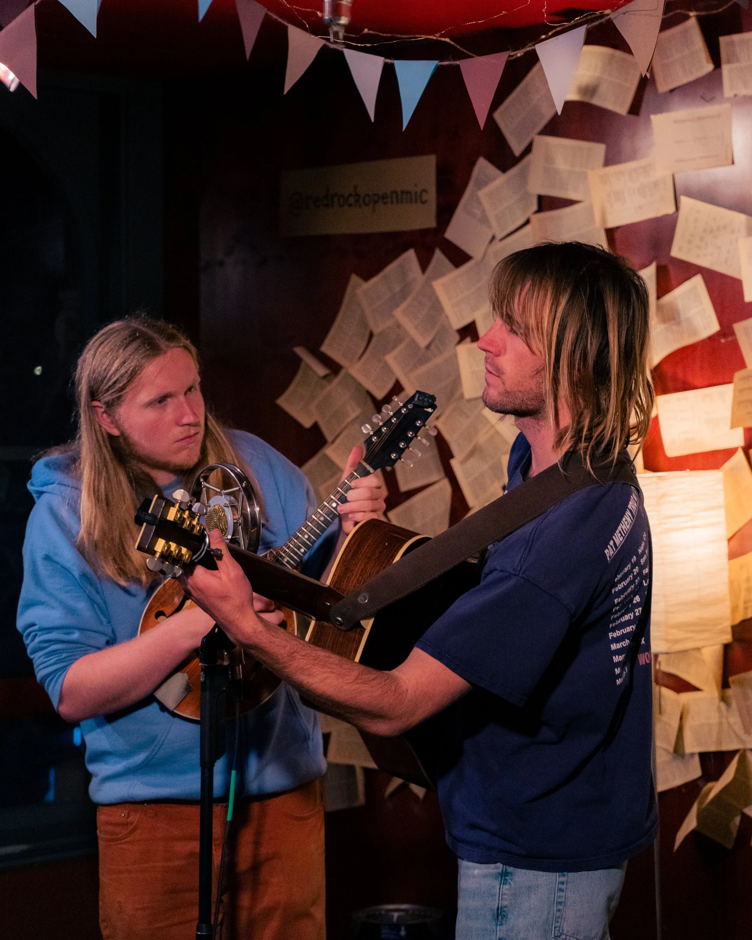 Two young men perform a folk music duet with acoustic guitars, in a dimly lit room decorated with papers and bunting banners.