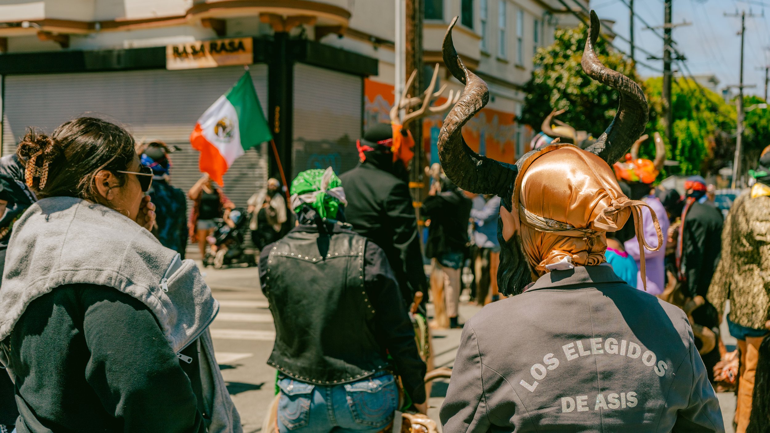 People participating in a parade or demonstration, with one person wearing a costume with large horns and a shiny gold and black headpiece, and others holding a Mexican flag.