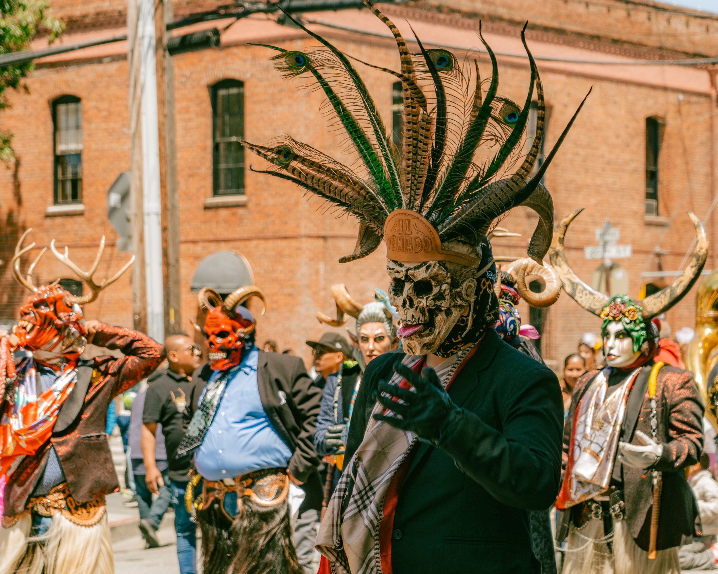 People dressed in elaborate costumes and masks with horned and feathered headdresses during a cultural event or parade on a city street.