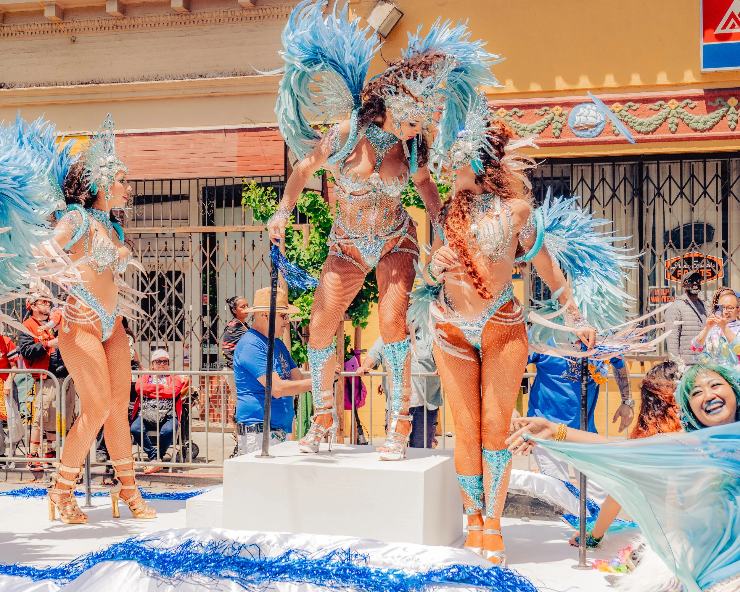 Women in colorful carnival costumes with feathers, rhinestones, and high heels, participating in a parade on a city street surrounded by spectators.