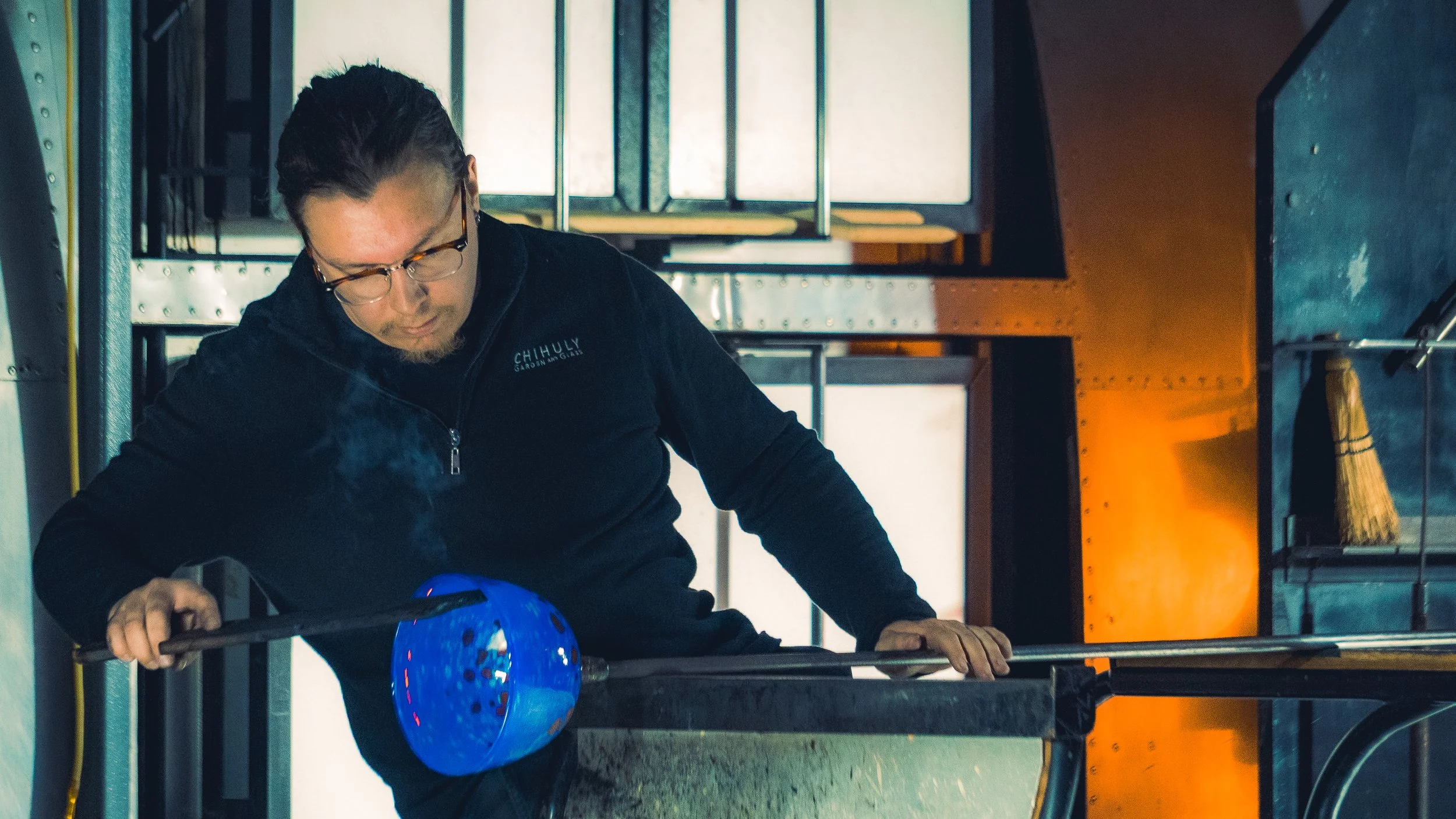A glassblowing artist shaping a blue molten glass piece on a blowpipe in a studio.
