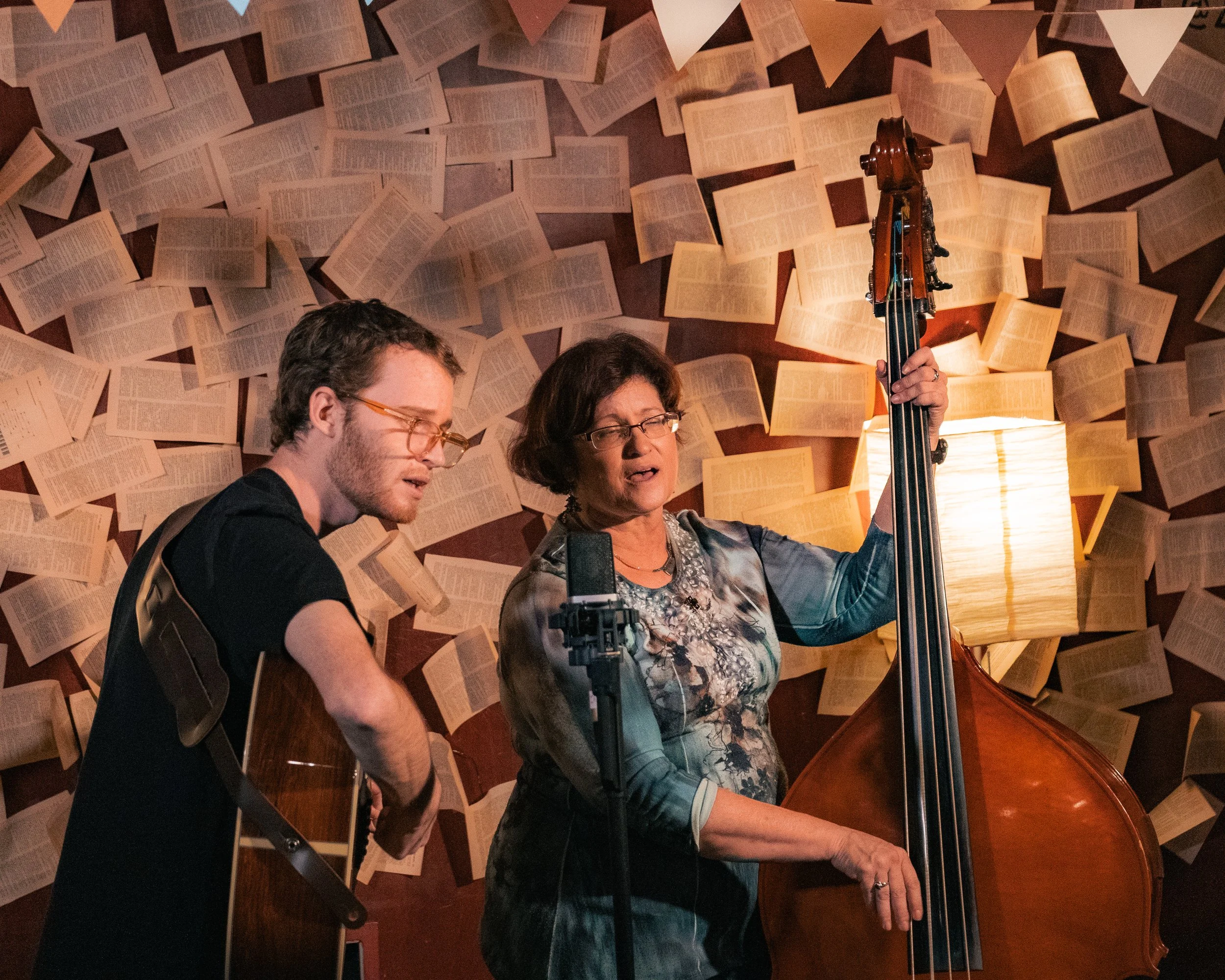 A woman playing a double bass and a young man playing an acoustic guitar, performing together in a dimly lit room decorated with open books on the wall and a lamp.