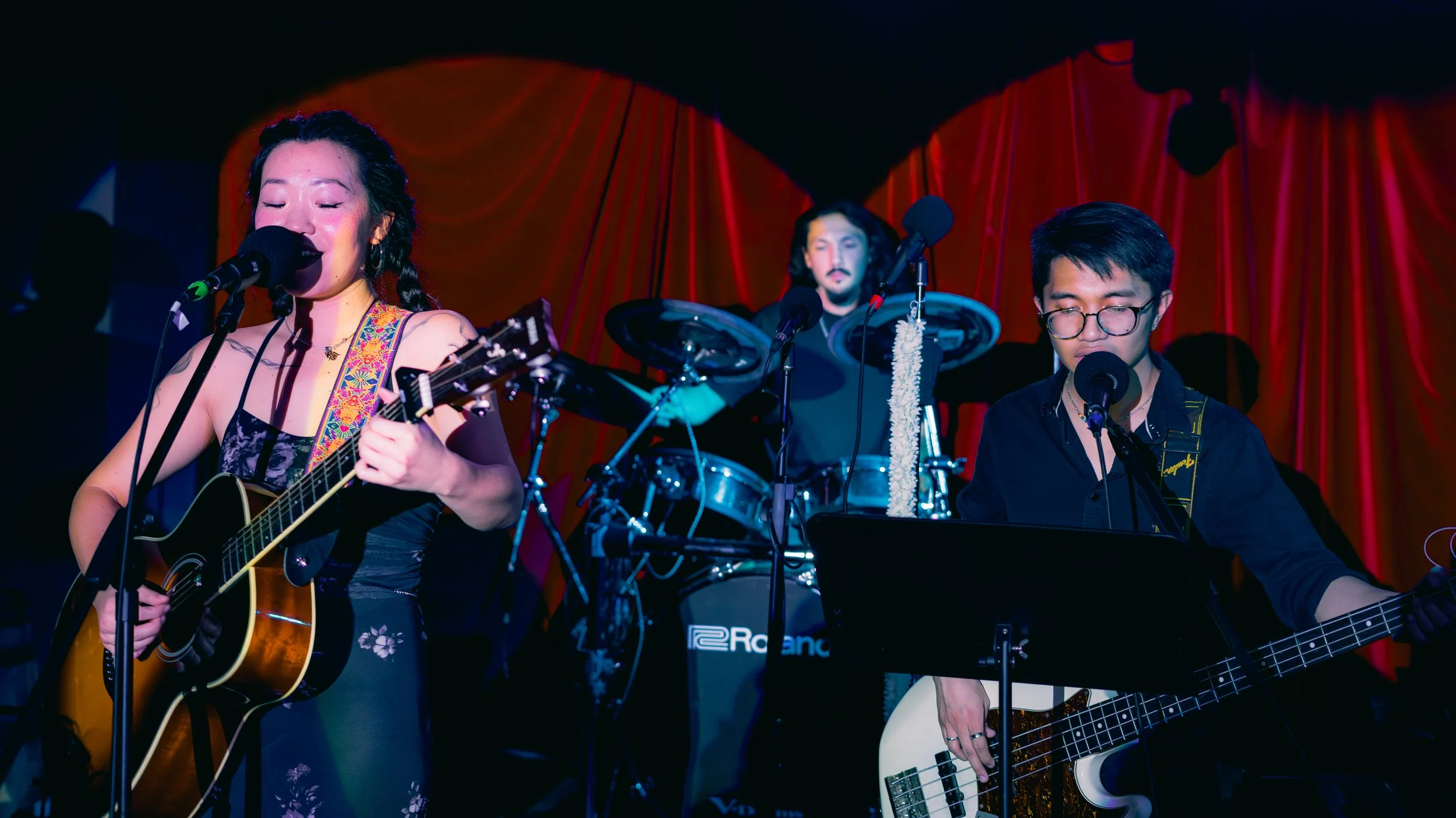 Three musicians performing on stage with a red curtain backdrop, including a woman playing an acoustic guitar and singing, a man playing a bass guitar, and a drummer behind them.