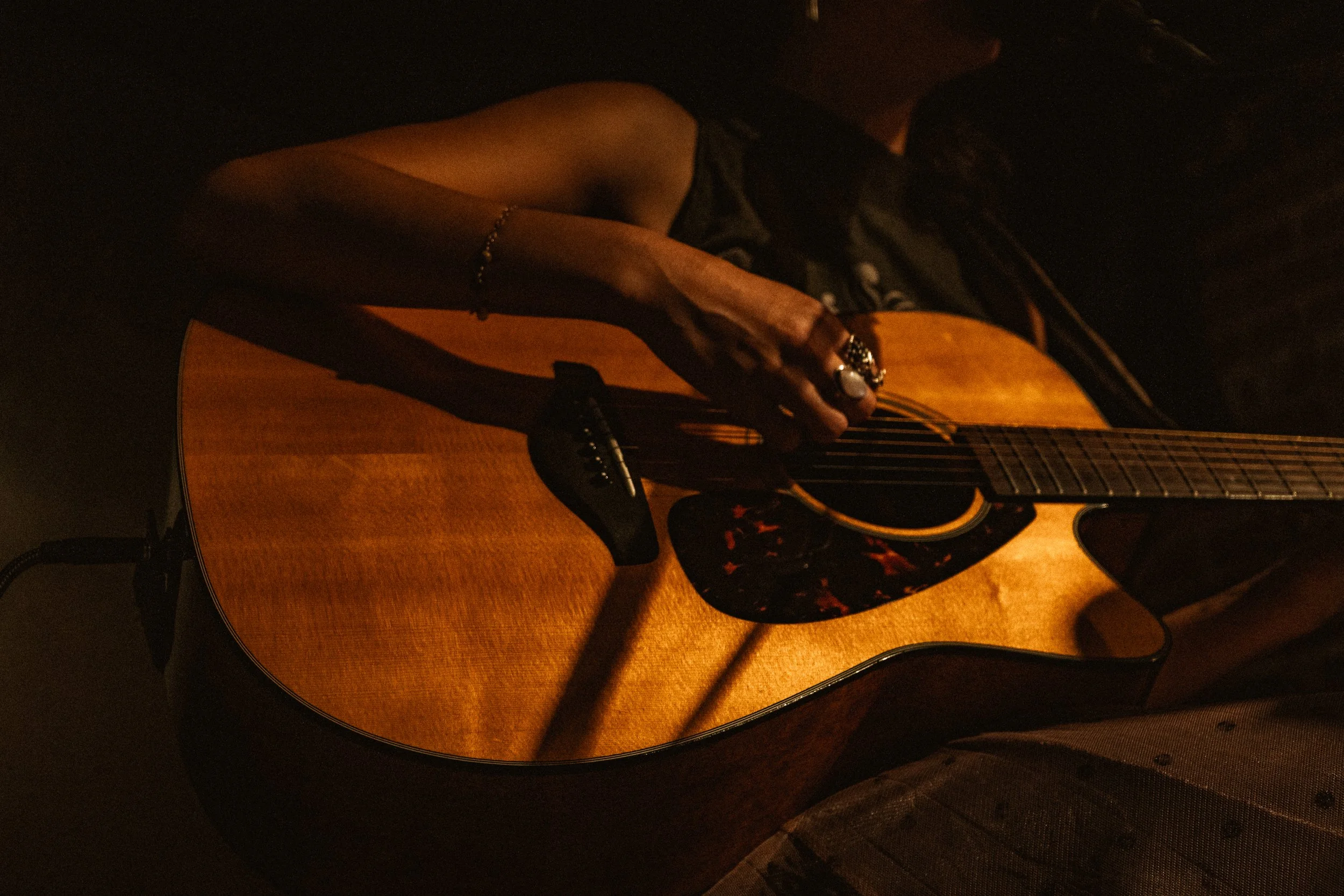 A person playing an acoustic guitar, partially illuminated in warm light, with a focus on their hand and the guitar's body.