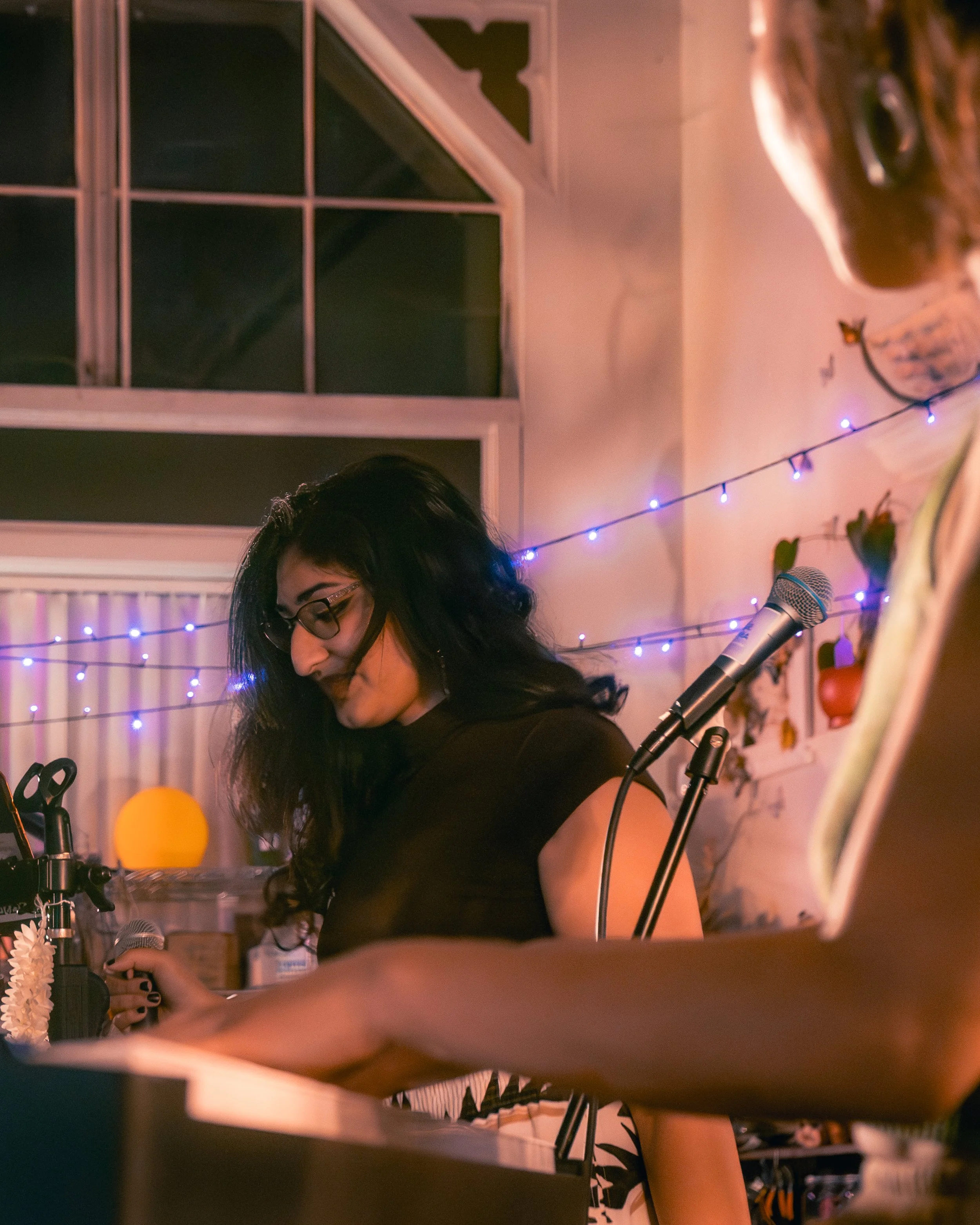 A woman with long black hair, glasses, and a black top standing at a microphone in a cozy room decorated with string lights and plants