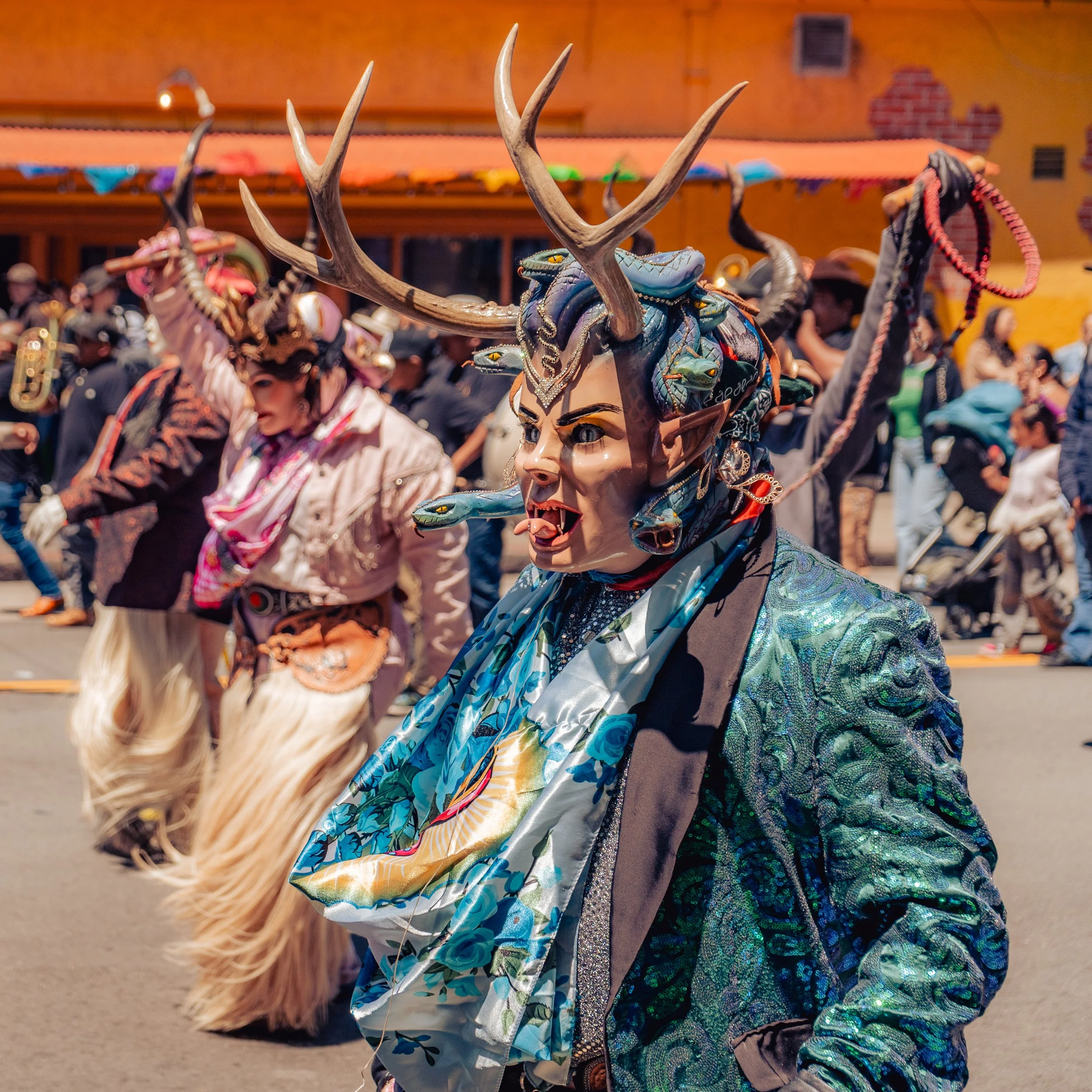 Person wearing an elaborate demon mask with large antlers, pointed ears, sharp teeth, and snake details, participating in a parade with dancers and musicians in the background.