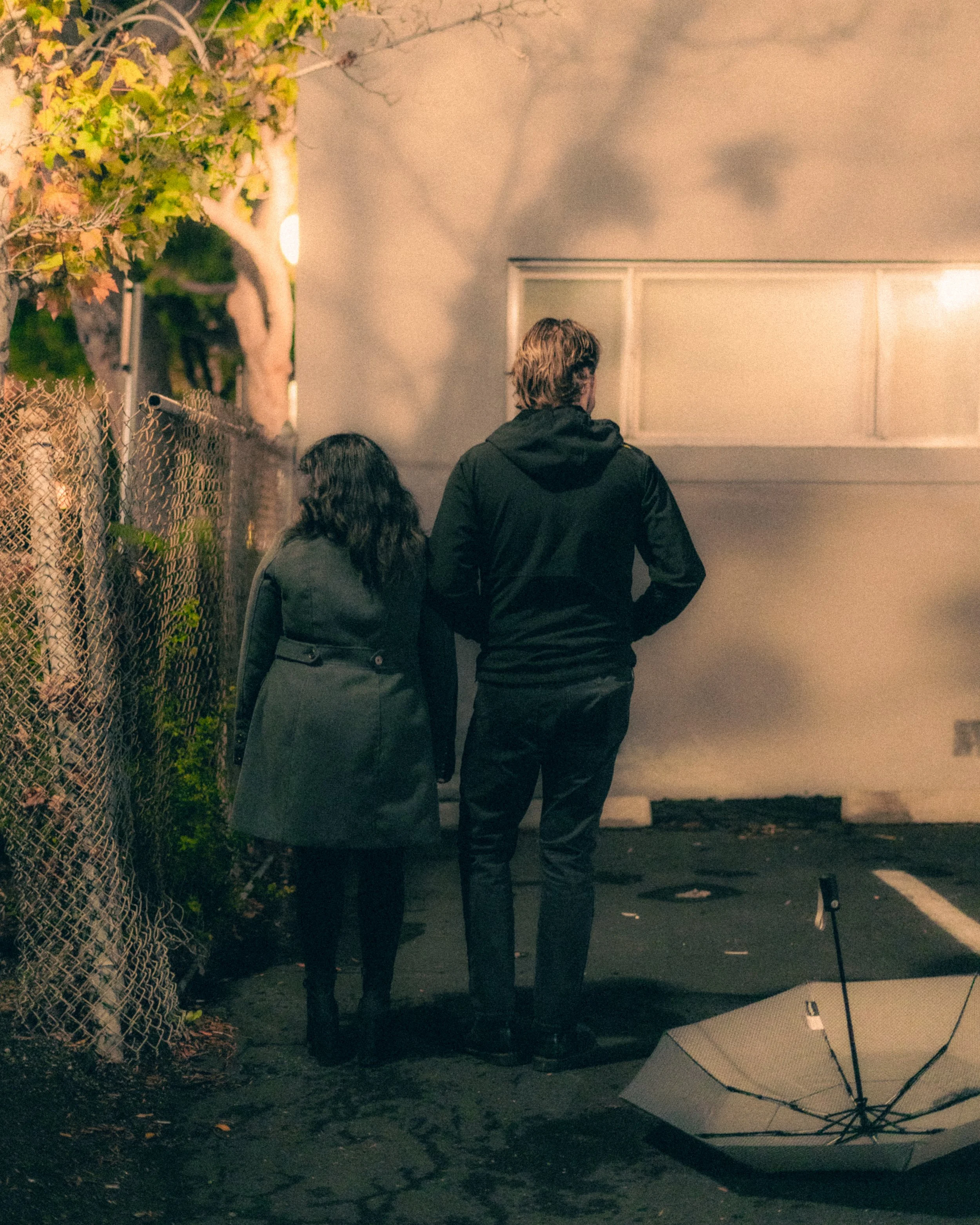 A man and woman walking together at night in an alley, with an umbrella on the ground nearby and a fence on the left side.