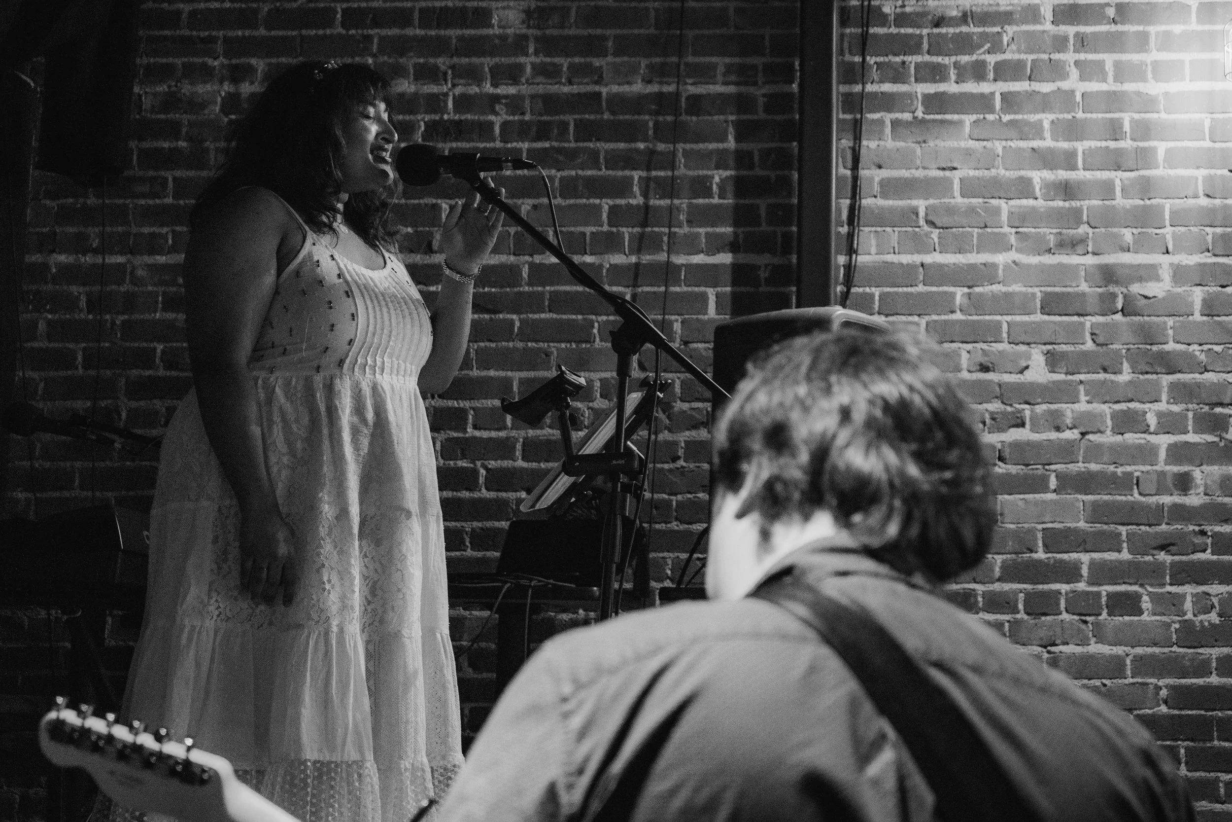 A woman sings into a microphone on stage with a brick wall background, while a man in the foreground listens, holding a guitar.