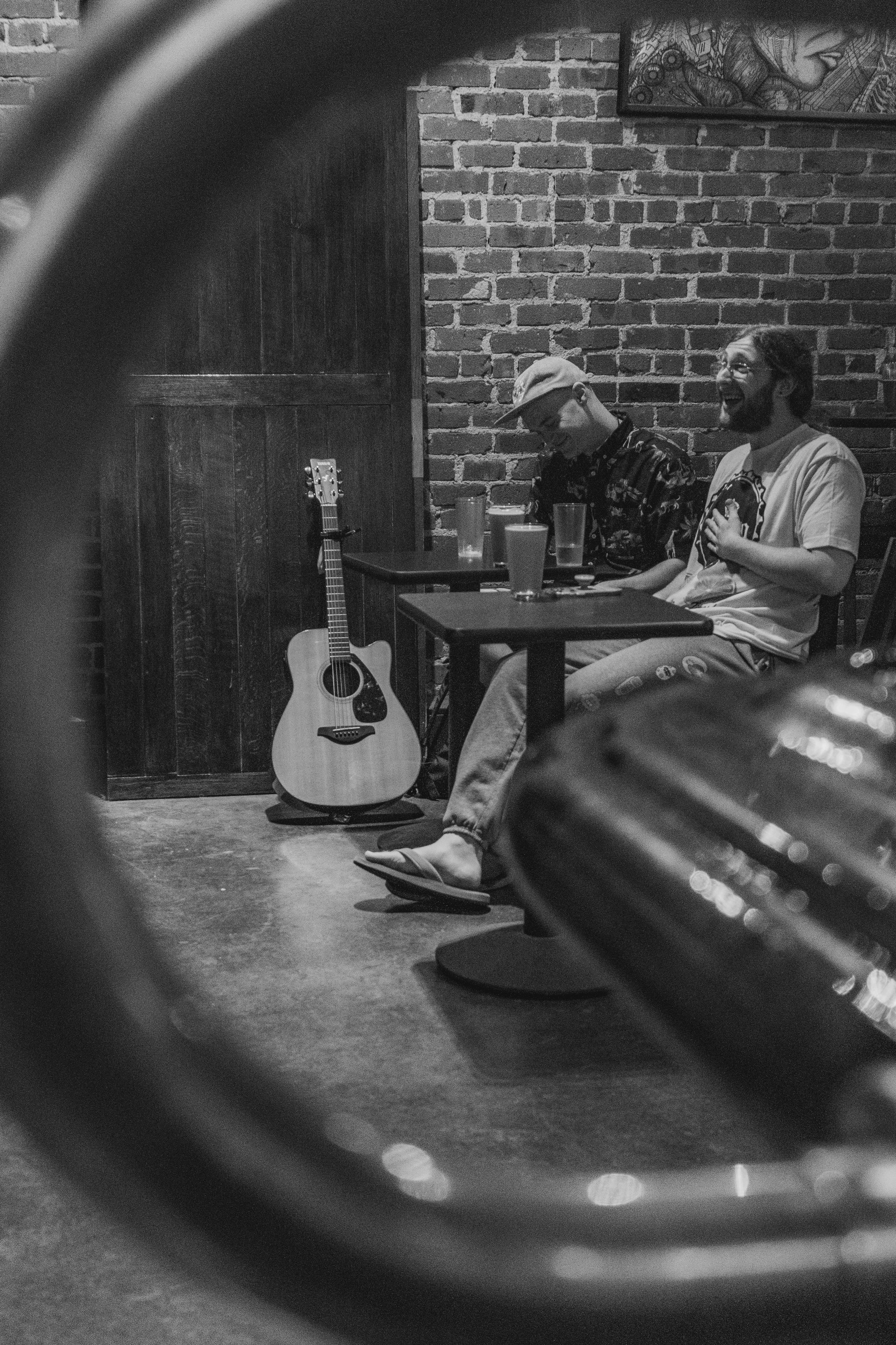 Two men sitting at a table in a brick-walled room, smiling and laughing, with a guitar leaning against the wall nearby.