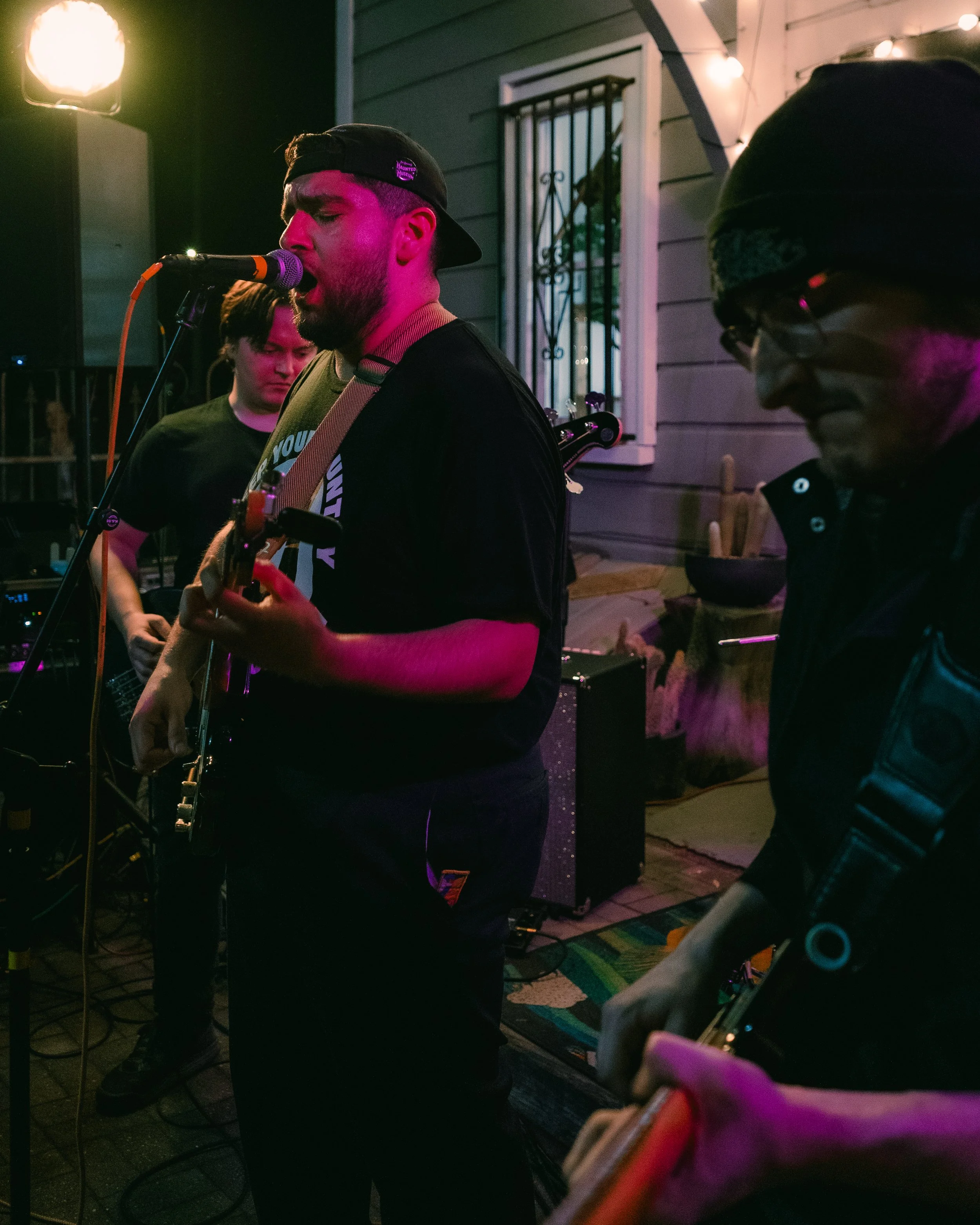 Three musicians performing with guitars and microphones on a porch, with string lights and house window visible.