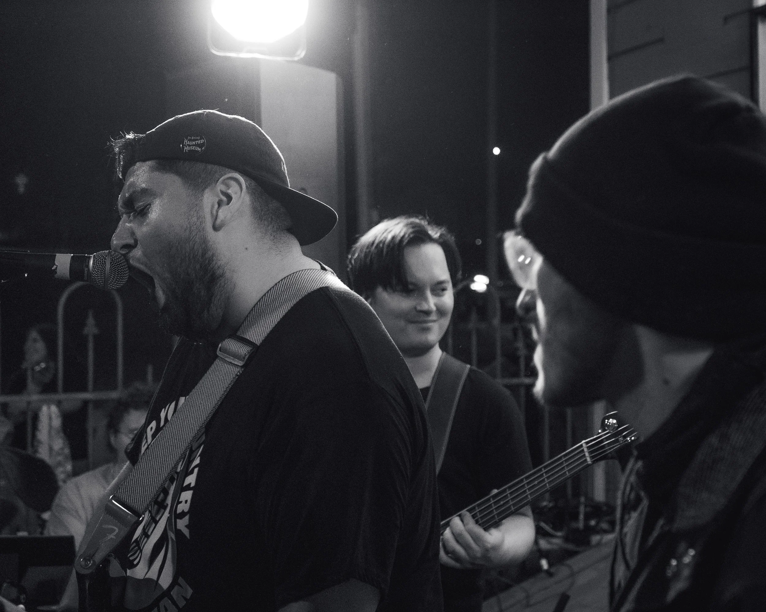 Three musicians performing outdoors at night, one singing into a microphone and wearing a cap, one smiling and holding a guitar, and another with glasses and a beanie.