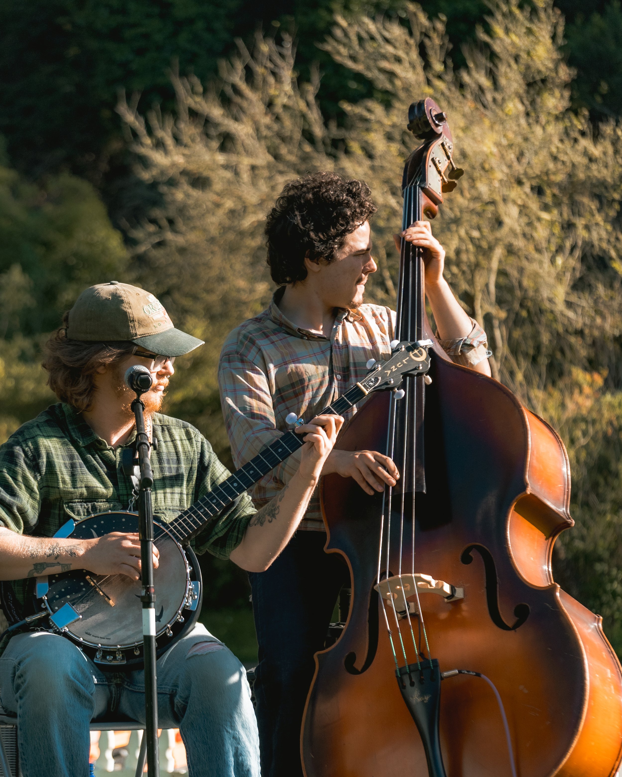 Two musicians playing string instruments outdoors during daytime, one with a banjo and the other with a double bass, with trees in the background.