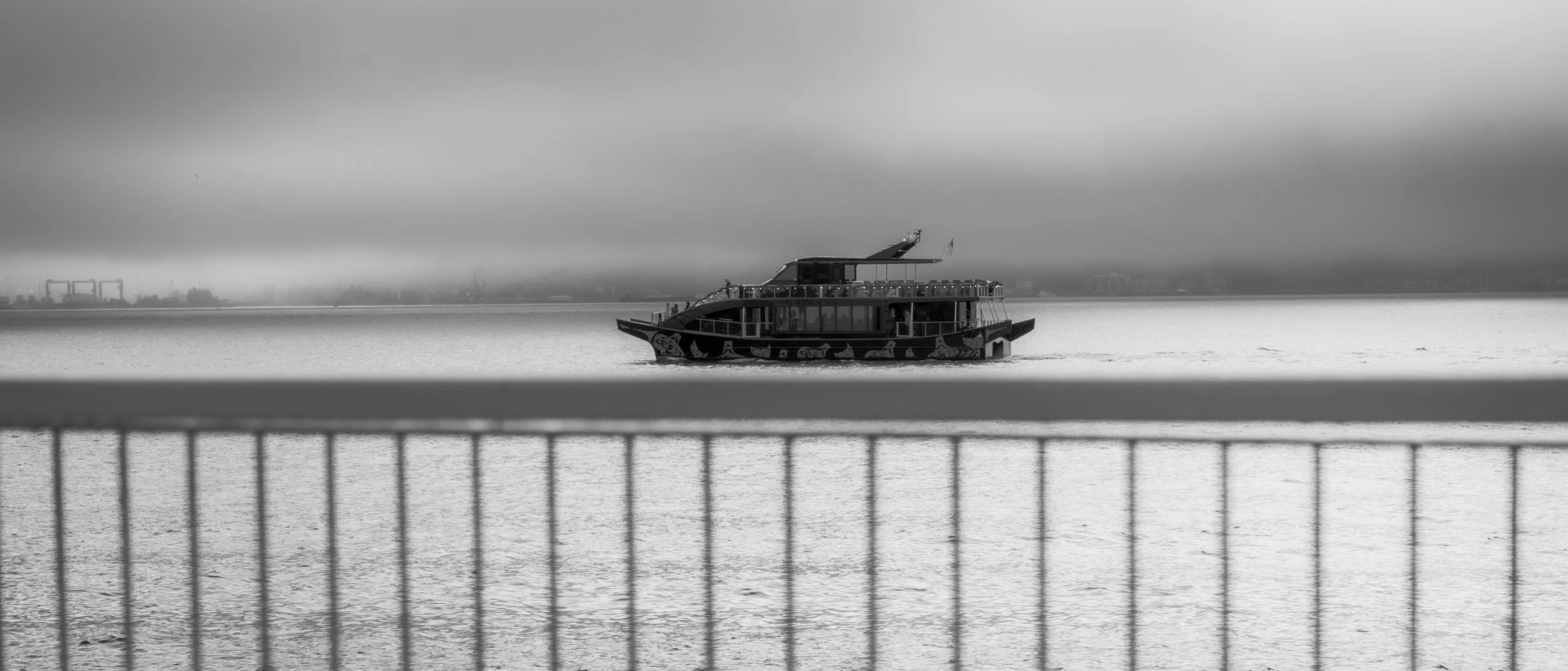 A black and white photo of a boat on calm water, taken from behind a railing. The boat has multiple decks and an American flag at the top. The sky is cloudy and foggy, with a faint cityscape and industrial structures in the distance across the water.