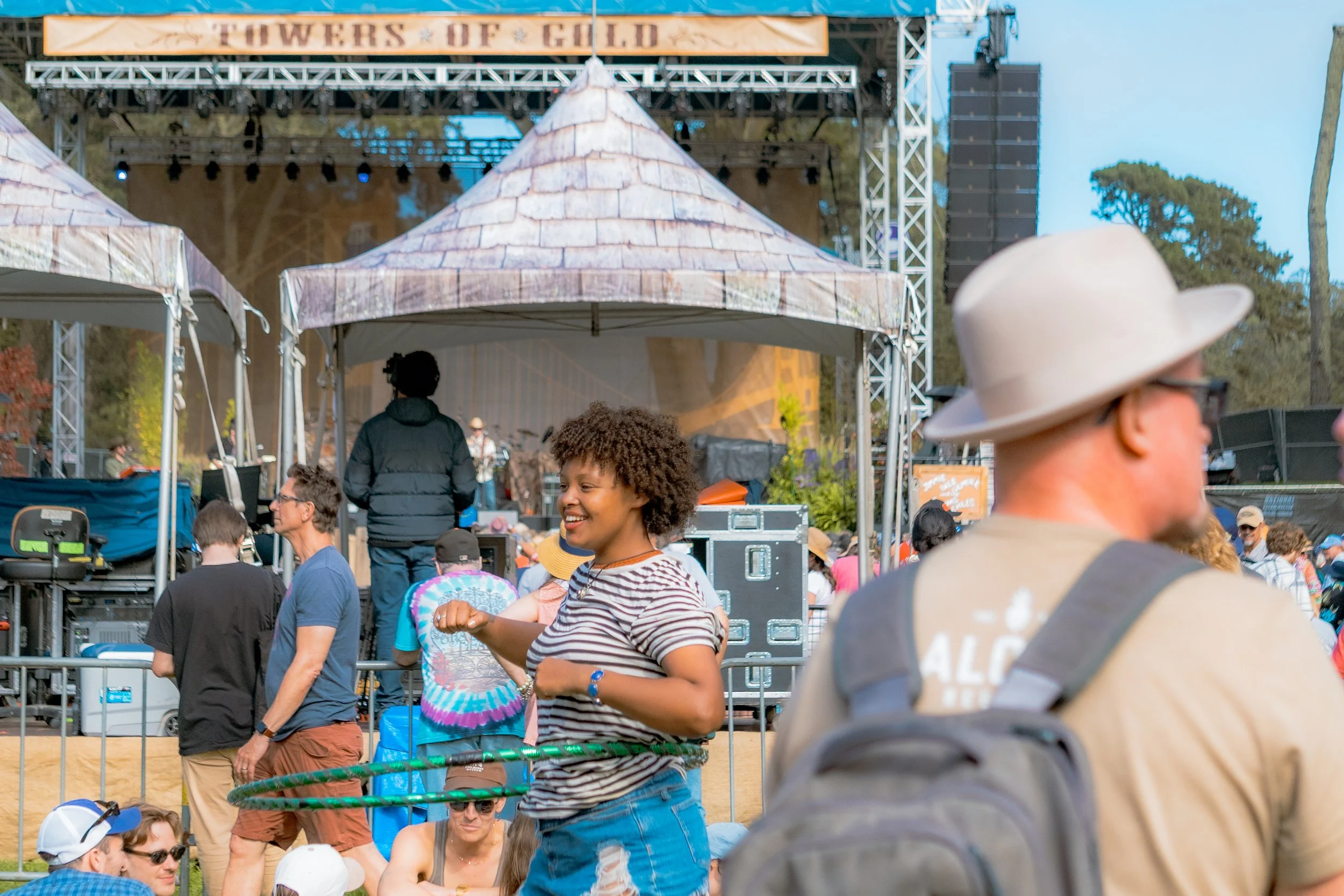 A woman with curly hair smiling and playing with a hula hoop at an outdoor music festival with a stage in the background, and several people standing or sitting around in casual clothing.