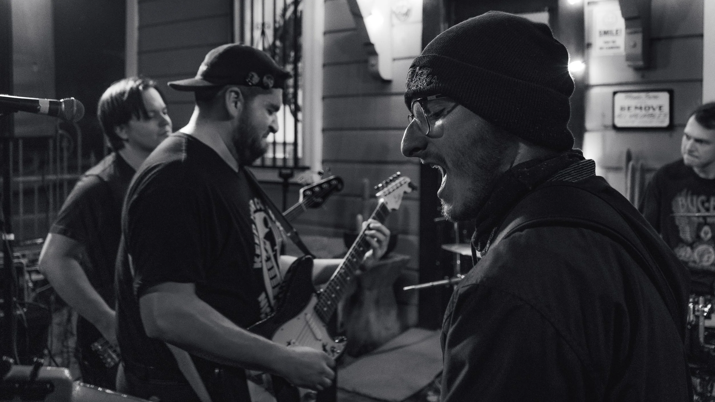 A group of four musicians performing outdoors at night, with one singing passionately, three playing guitars, and a drum set partially visible, all depicted in black and white.