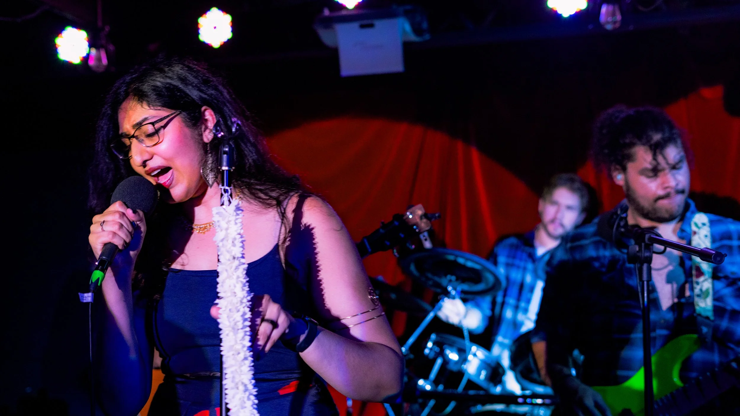 A woman with long black hair and glasses singing into a microphone on stage, accompanied by two men playing guitar and drums in the background, with colorful stage lighting and red curtains.