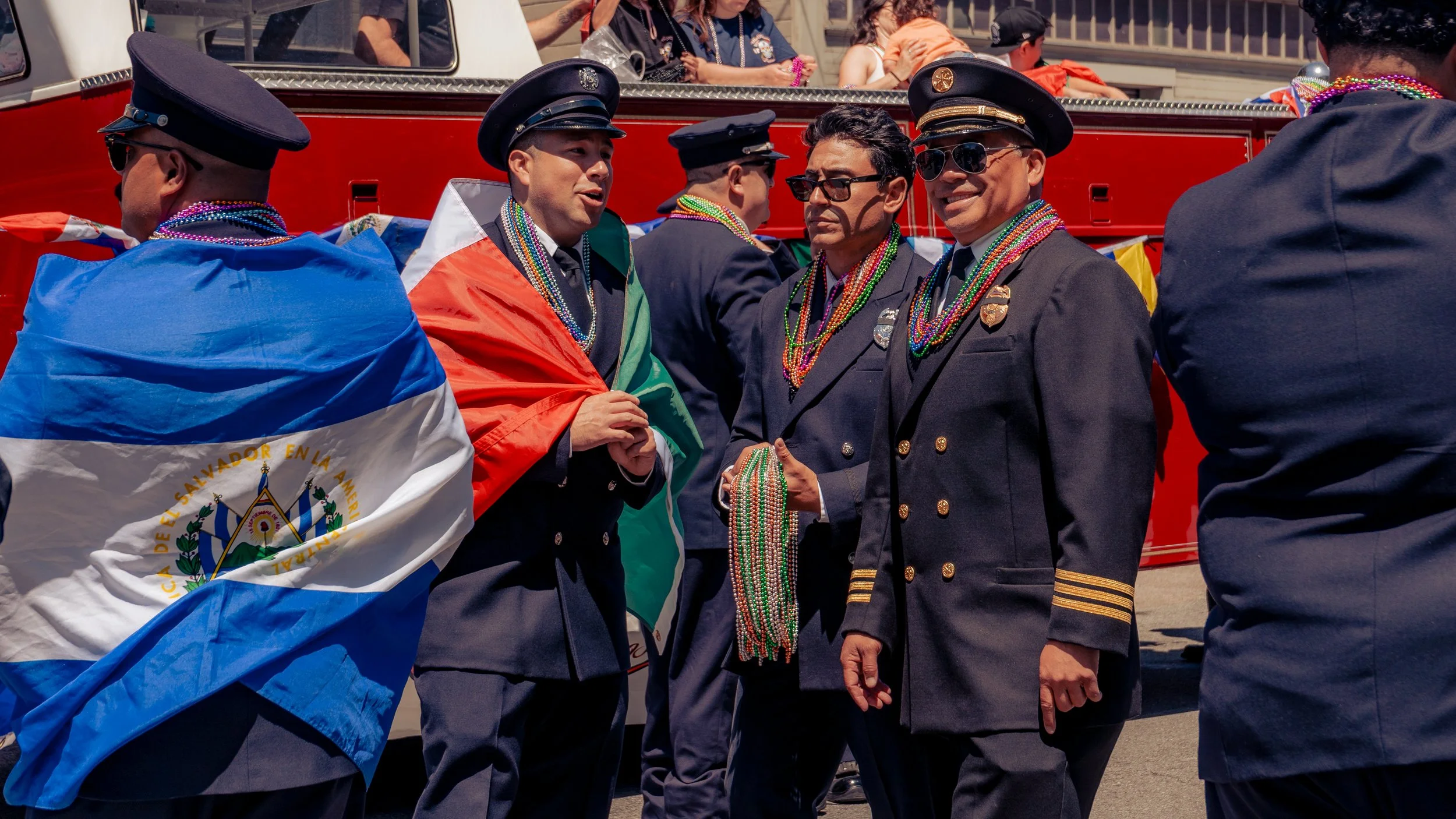 Men in uniform celebrating in a parade, with some wearing neck beads and one with a Salvadoran flag draped over his shoulders.