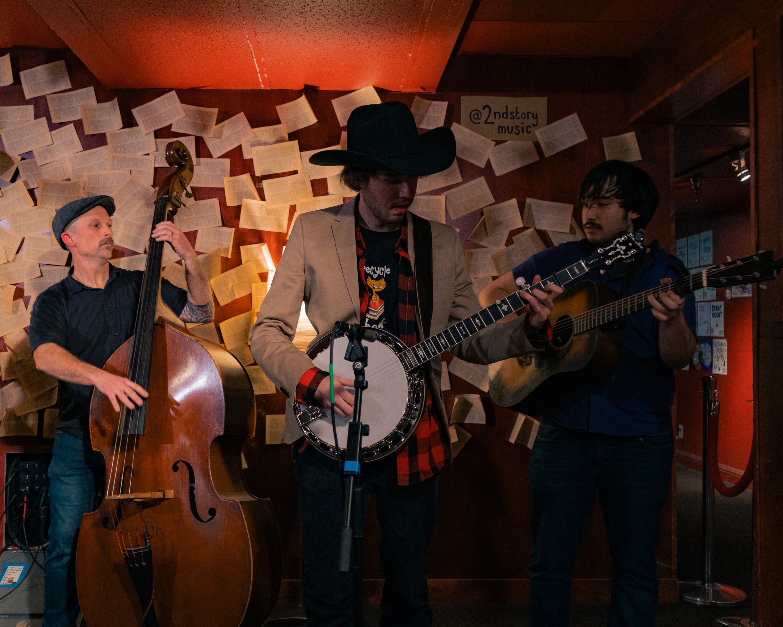 Three musicians performing indoors, with one playing upright bass, one playing banjo, and one playing acoustic guitar, against a background decorated with open books on the wall.