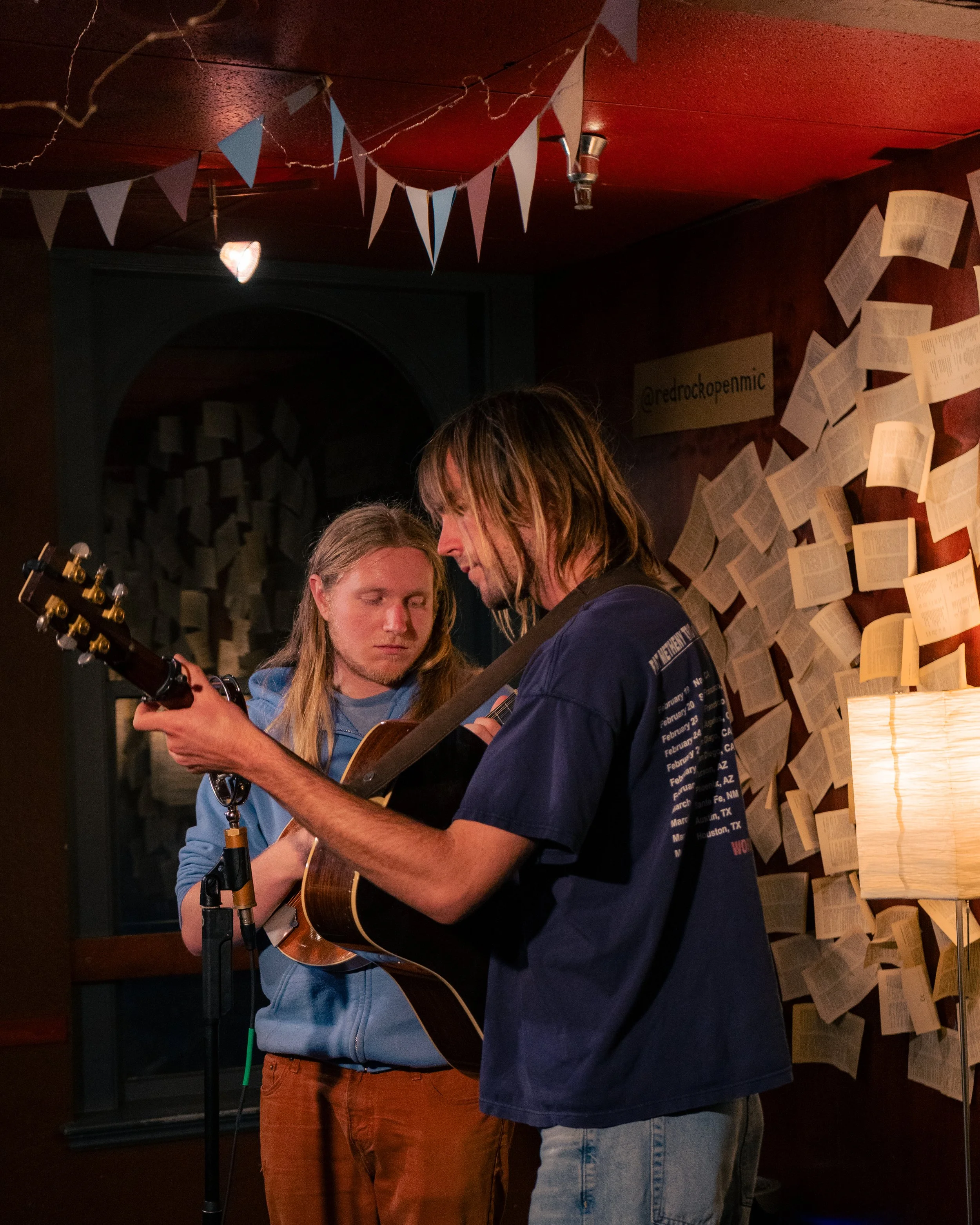 Two young men with long hair are performing with guitars in a dimly lit room decorated with numerous open pages of books on the wall and triangular bunting hanging from the ceiling.