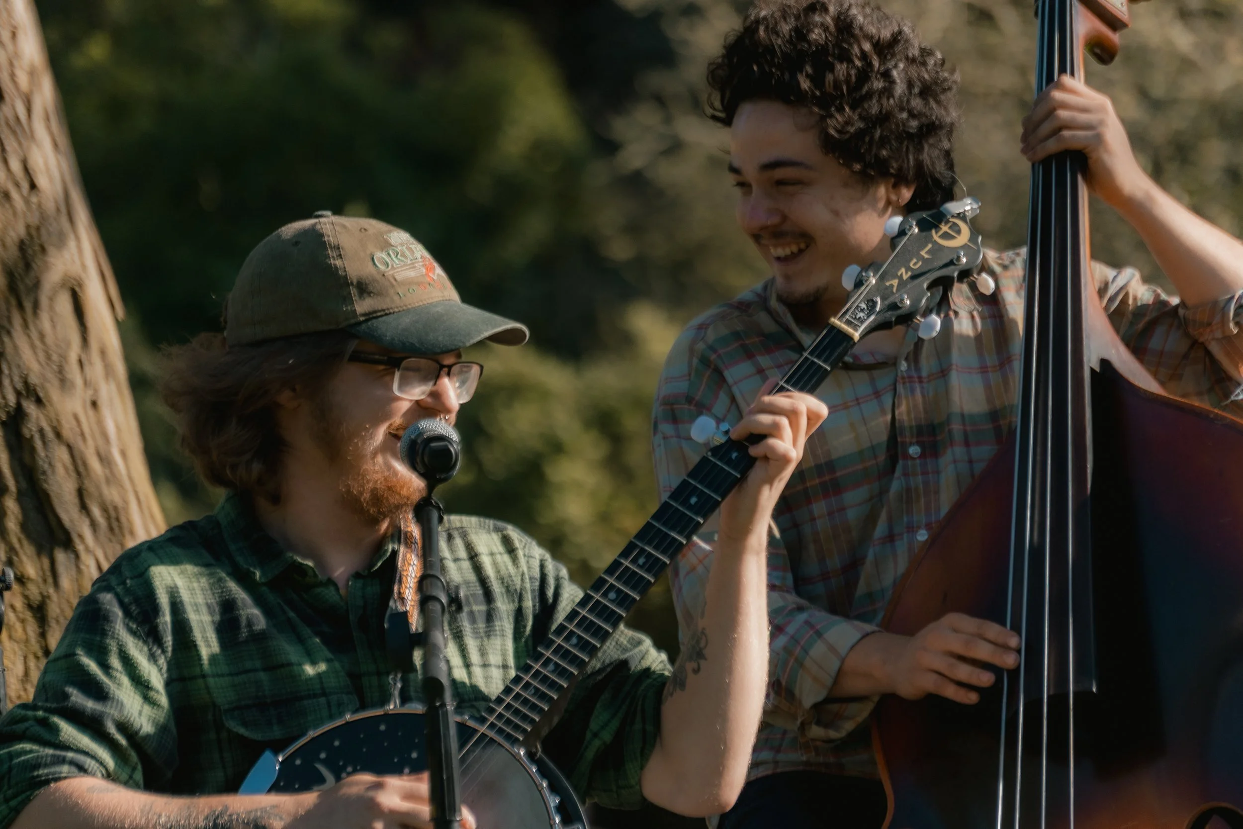 Two men playing string instruments outdoors, smiling and enjoying music, with a background of trees.