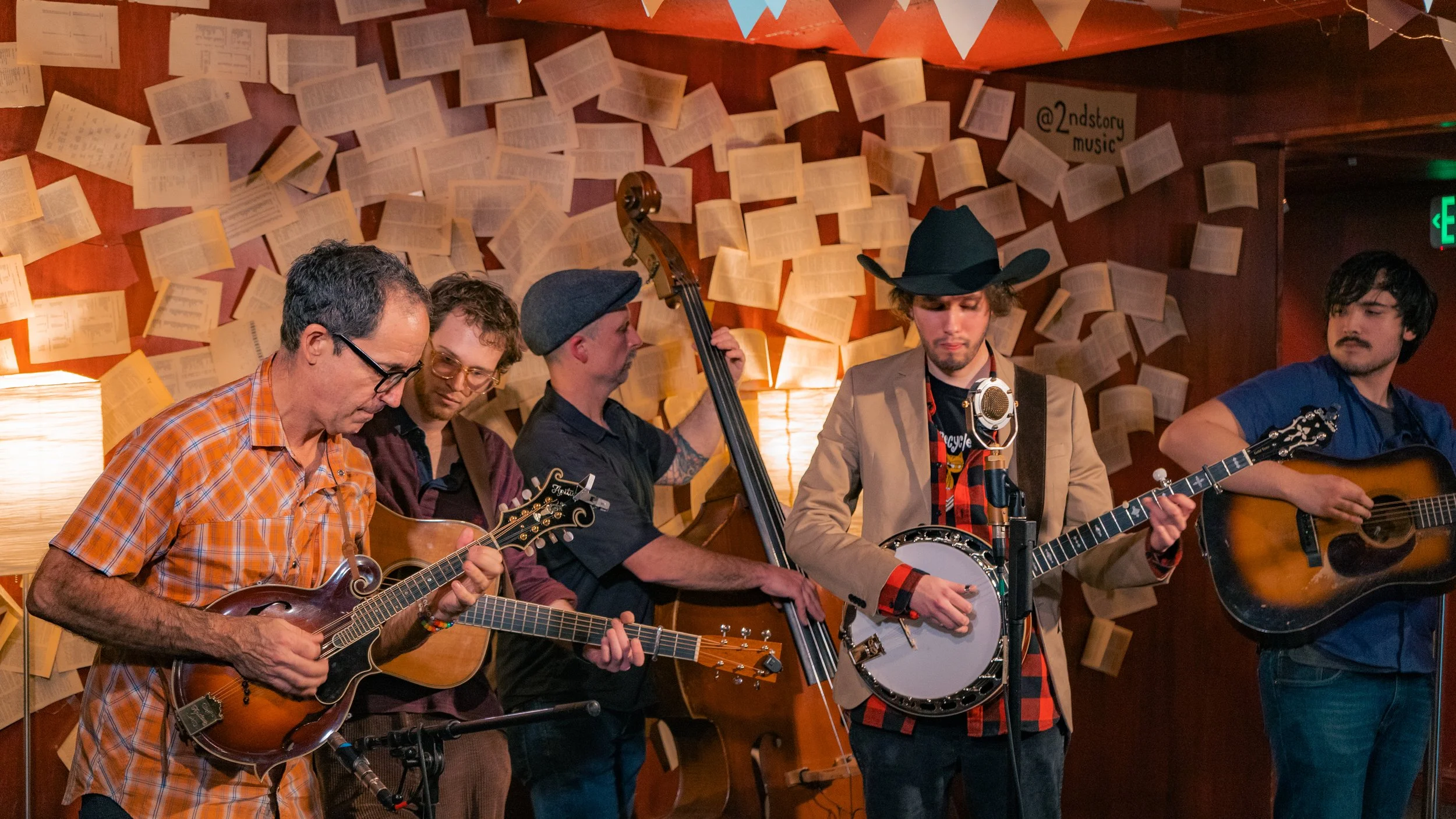 Five musicians playing instruments on stage, with open books or sheets on the wall behind them, illuminated by warm lighting.